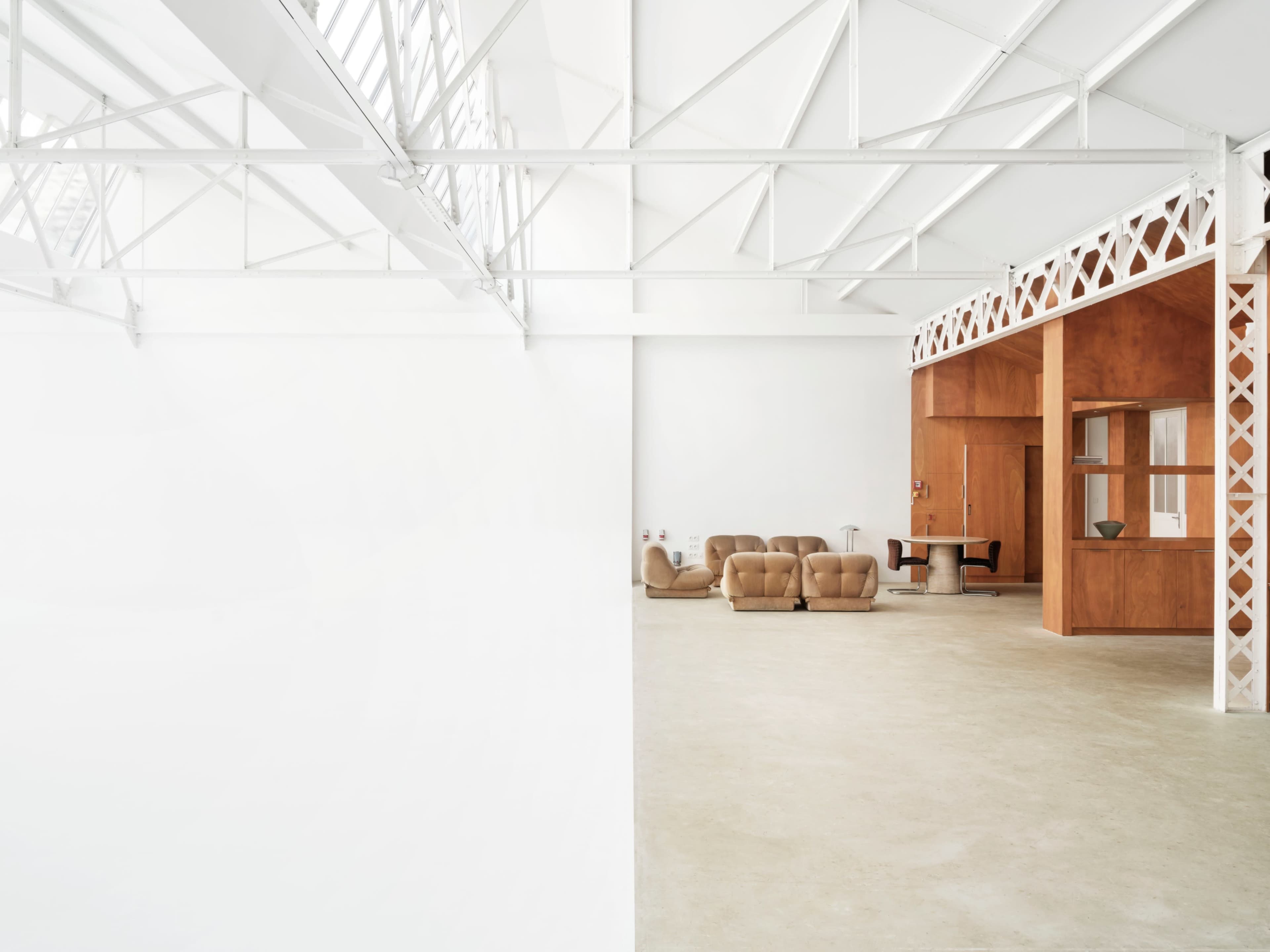 The image shows a spacious, minimalist interior with light-colored walls, a tan sofa, and wooden furniture.