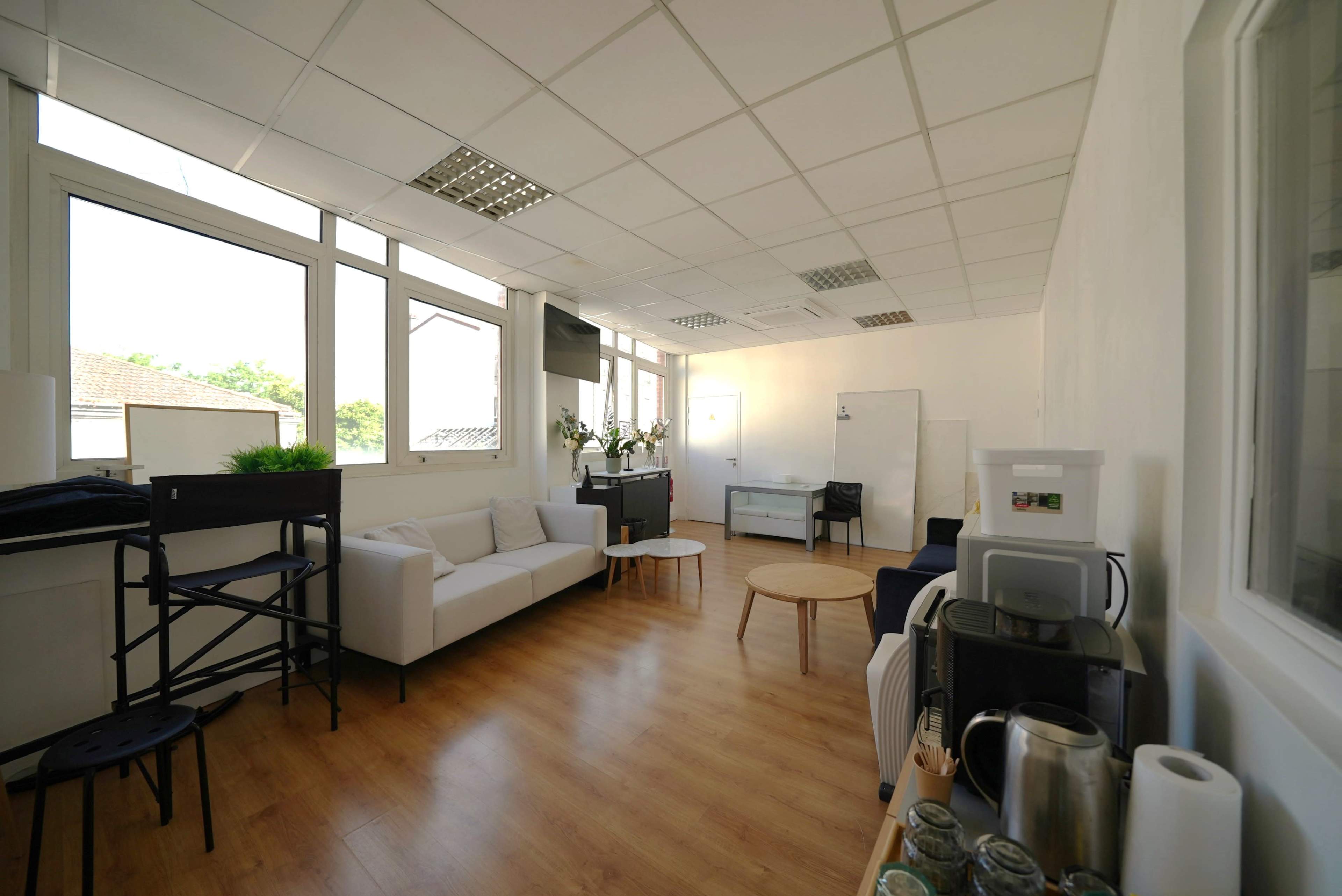 The image shows a spacious, well-lit office lounge area with a sofa, a coffee station, a small round table, and large windows along one wall.