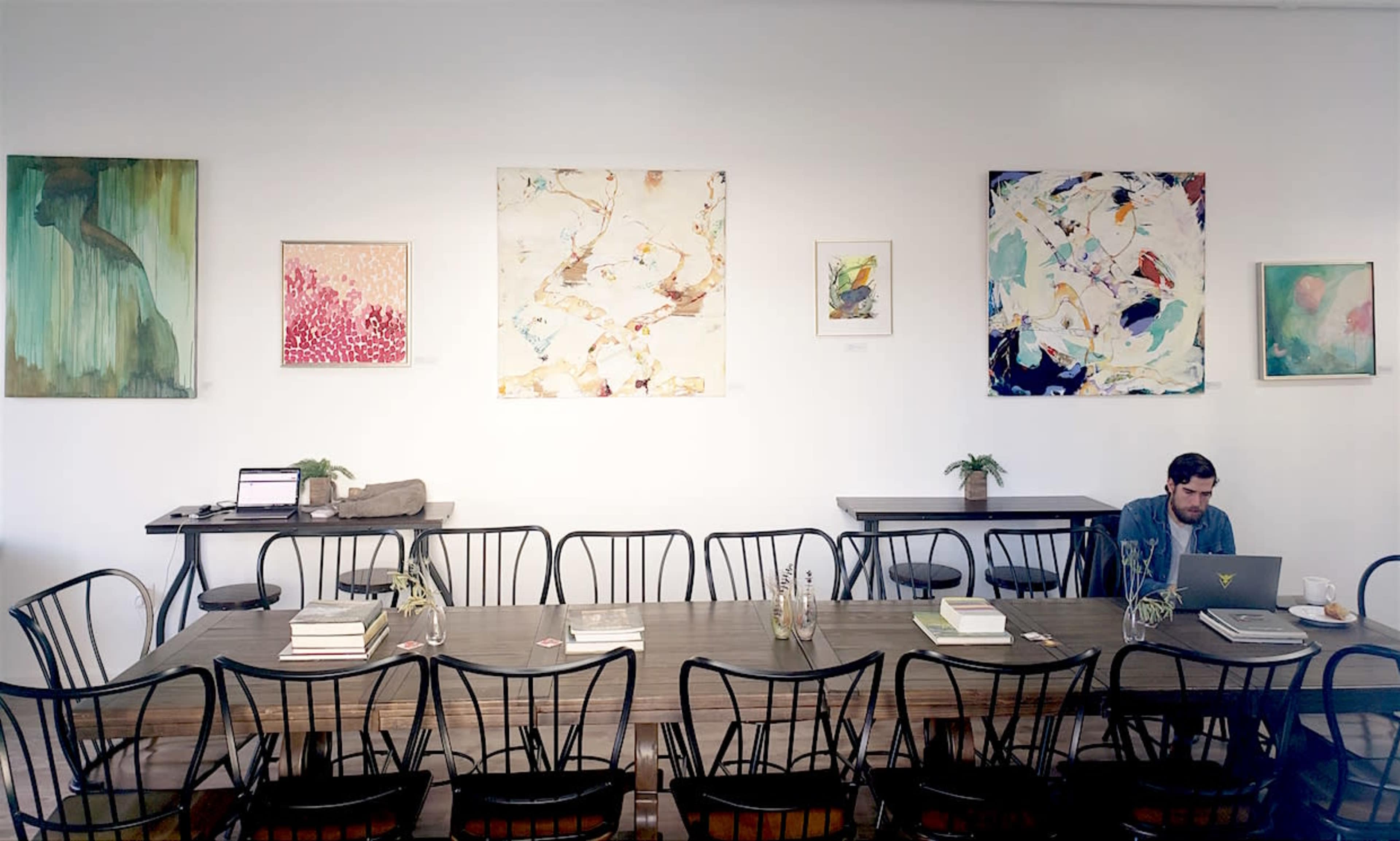 A long wooden table with black chairs occupies the center of a bright room adorned with various pieces of abstract art on the white walls.