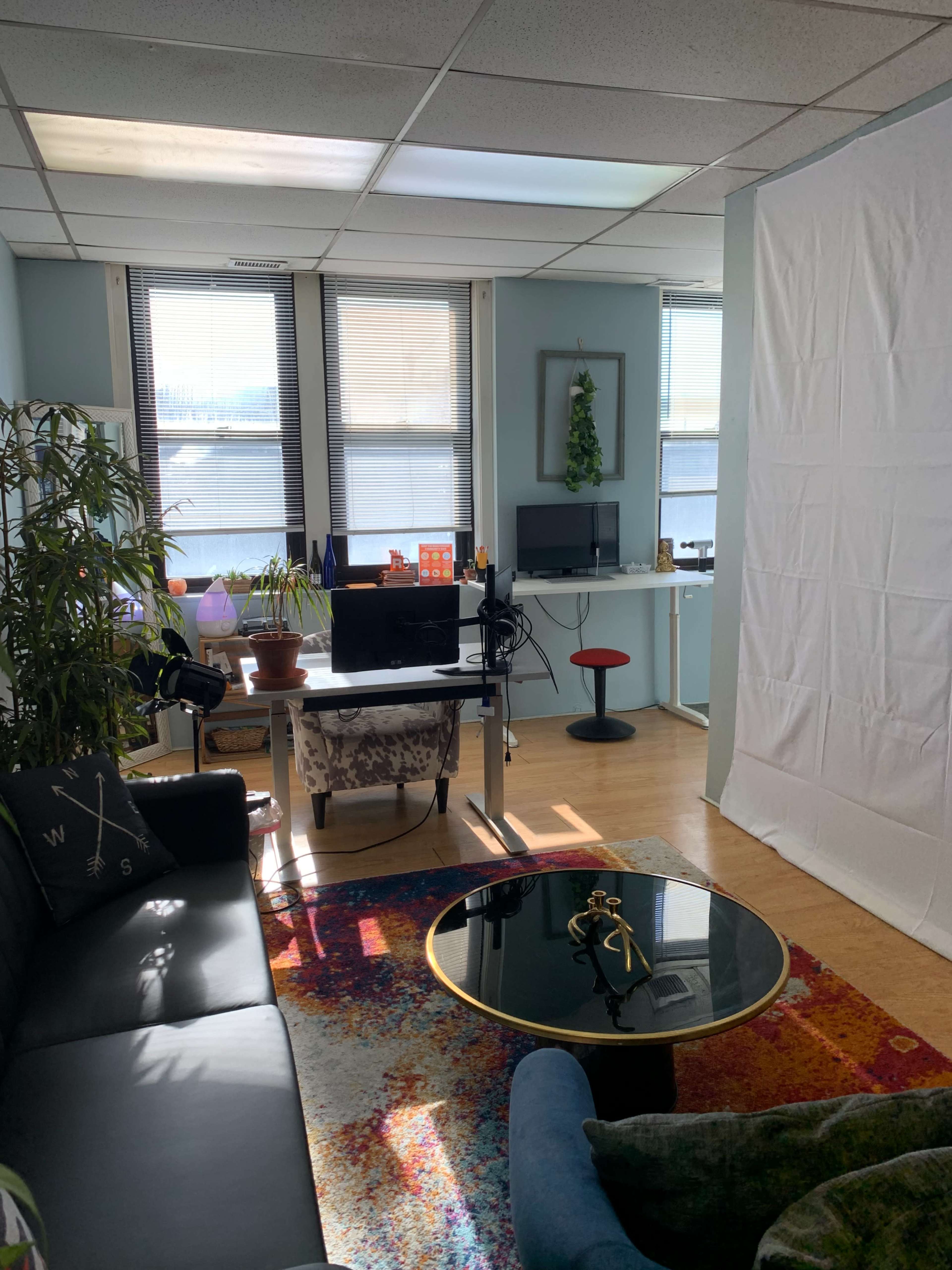 A well-lit office space with a couch, a coffee table, a desk with a monitor, and a backdrop setup in one corner.