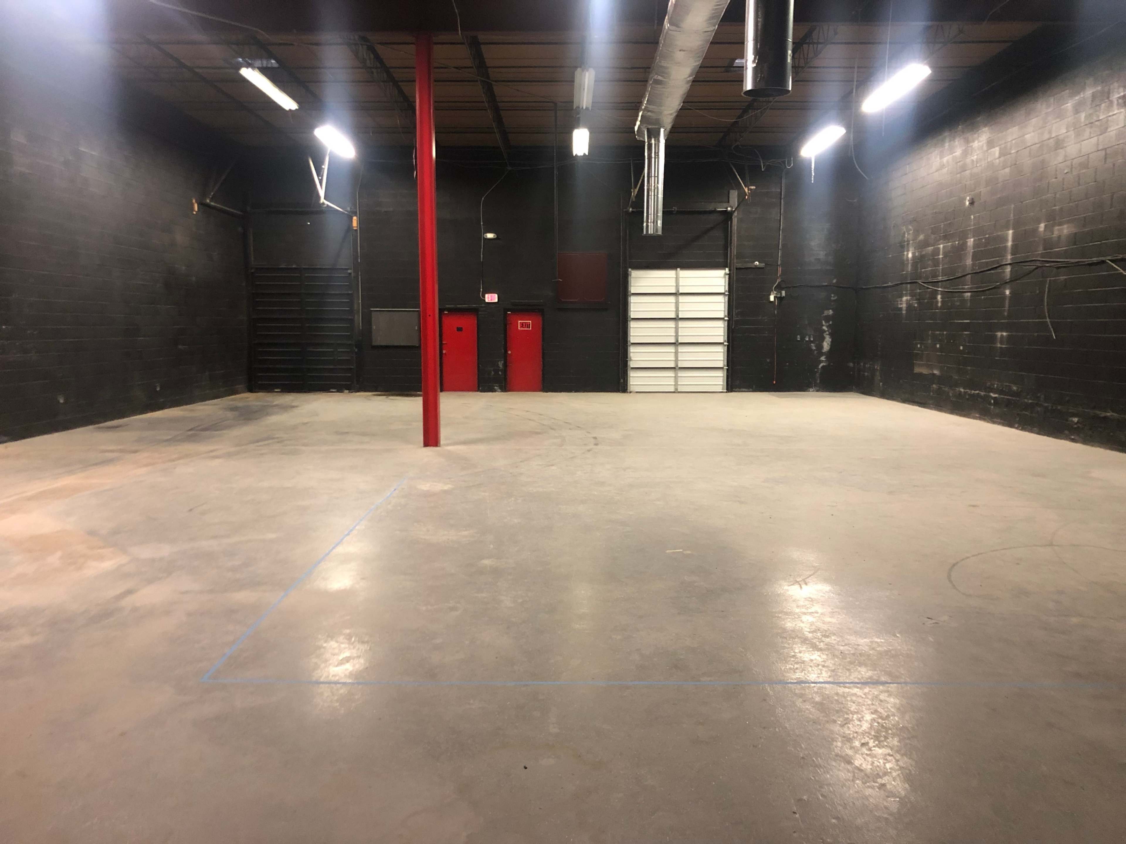 A large, empty industrial space with gray concrete flooring, black walls, and a central red support column.
