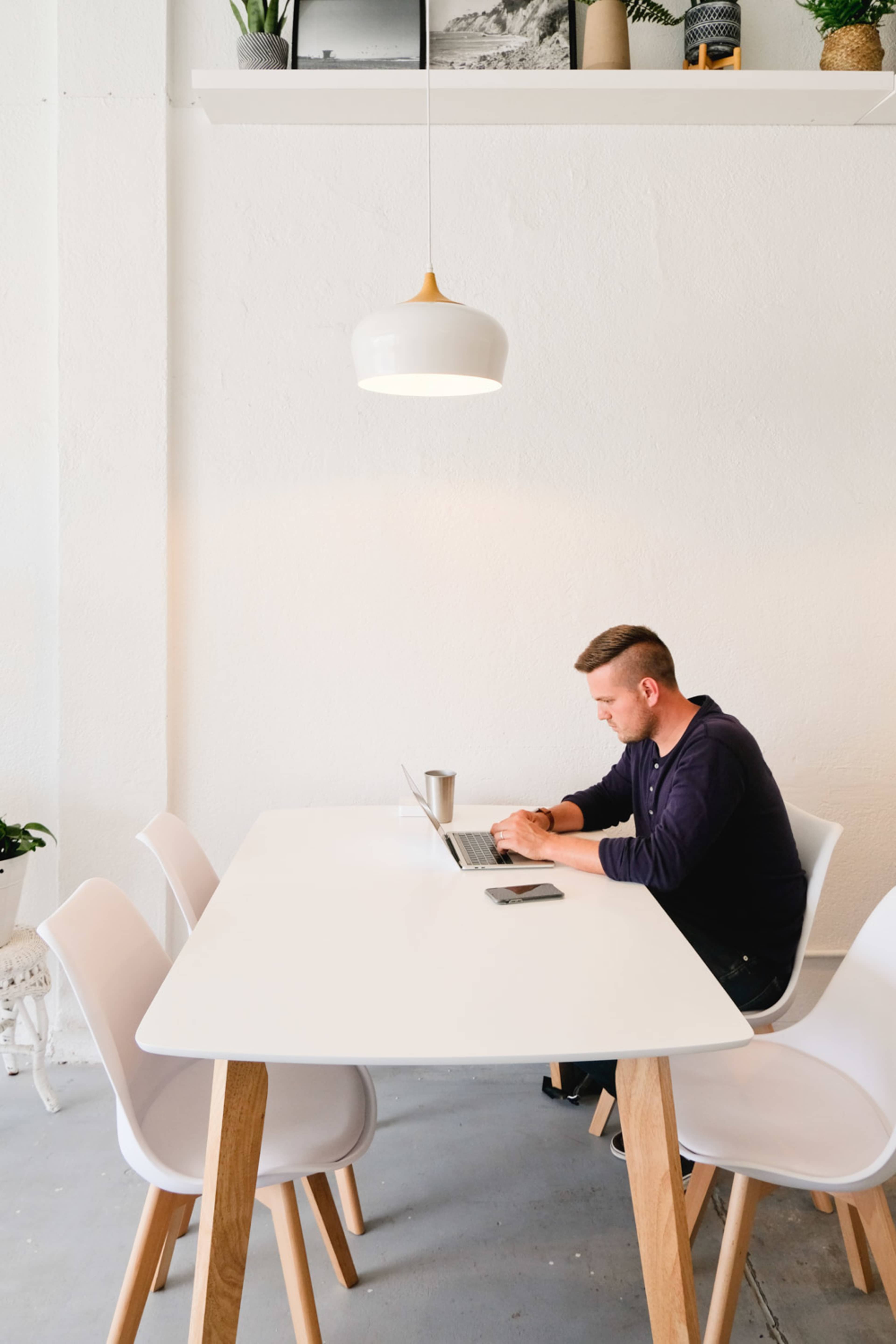 A man sits at a white table working on a laptop in a well-lit, minimalist room with wooden accents and a few plants.