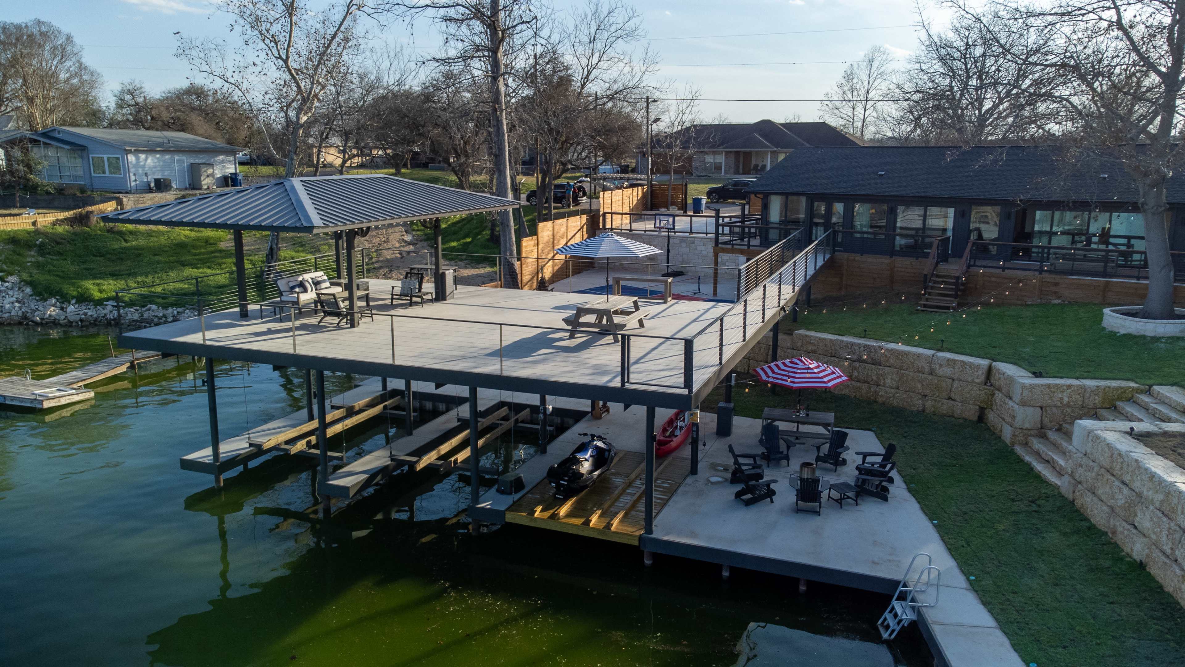 The image shows a large wooden dock with seating areas and a covered pavilion on a lake, surrounded by residential houses and trees.