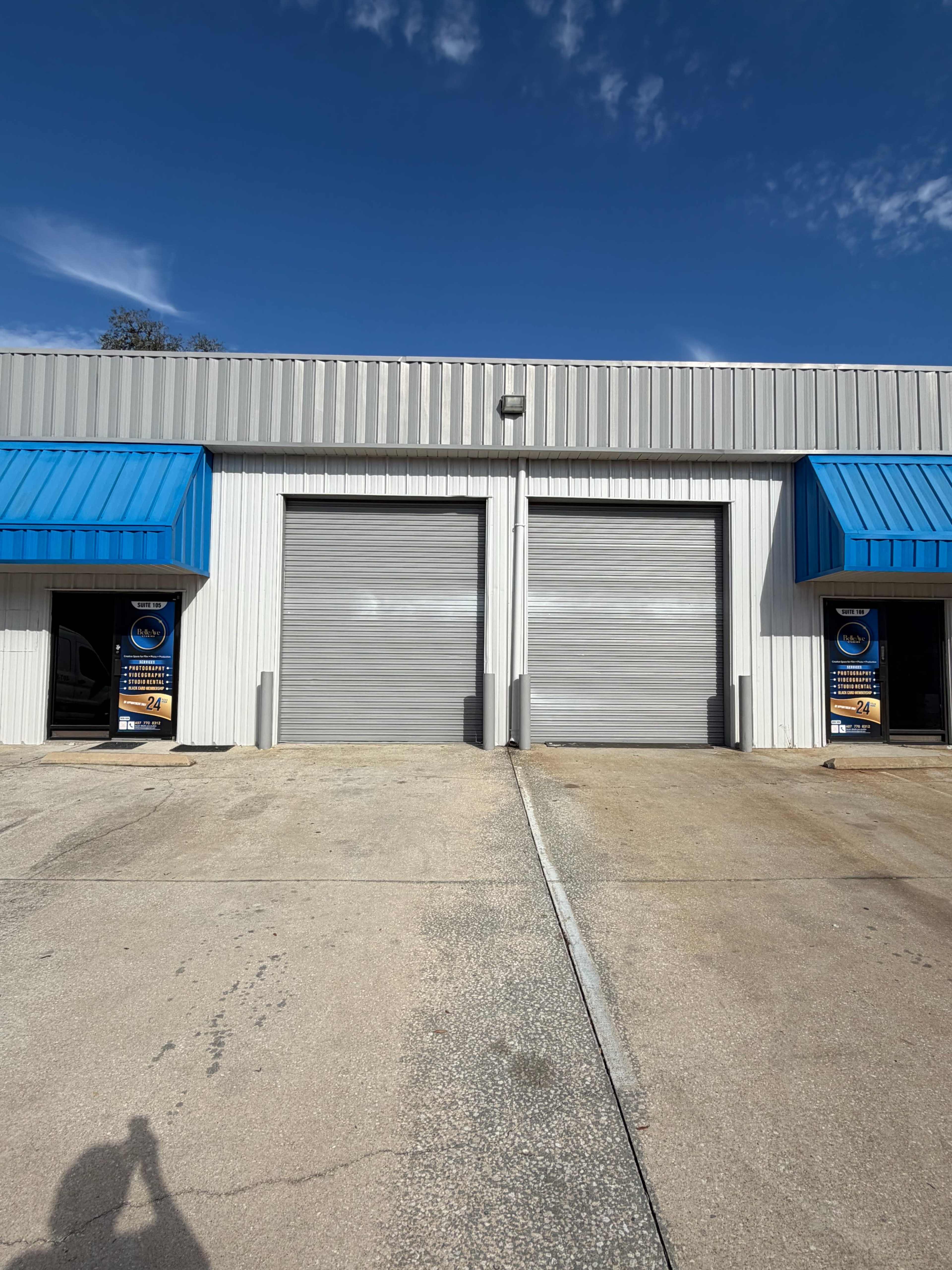 The image shows a commercial building with two closed metal roll-up doors, flanked by blue awnings and promotional banners near the entrance.