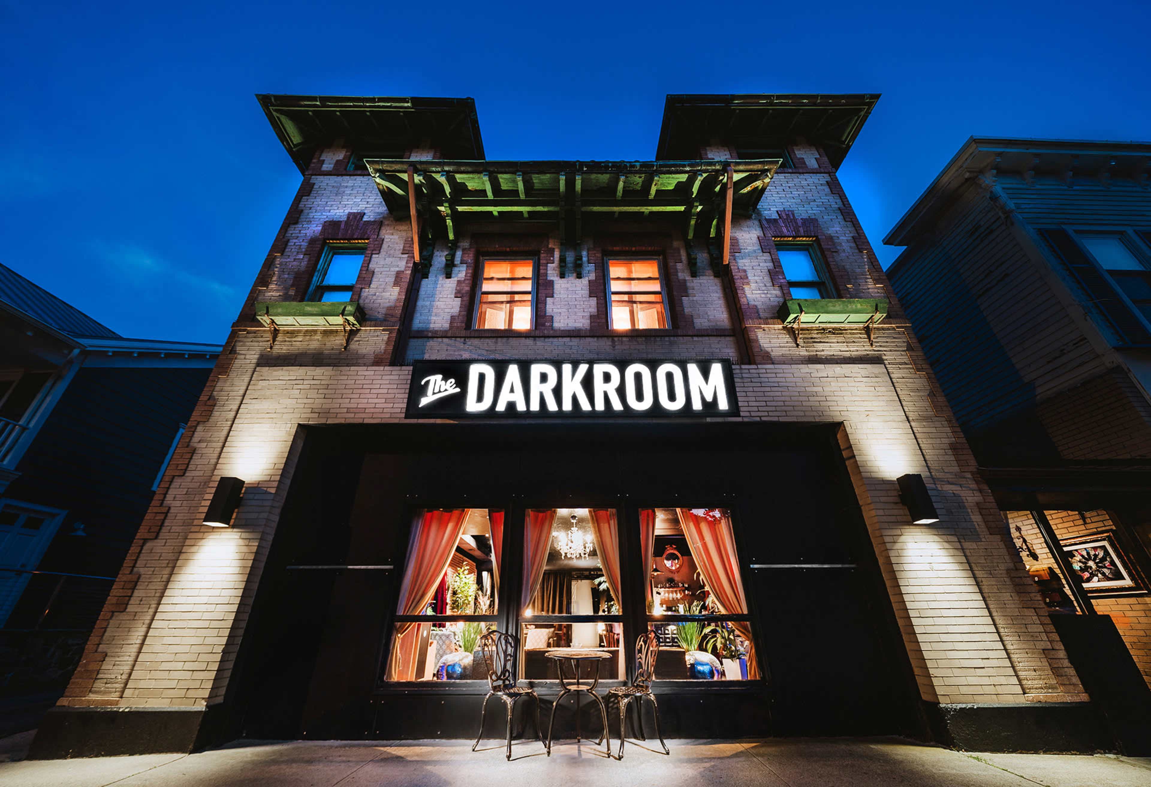 The image shows a building with a sign reading "The DARKROOM" and large windows framed by red curtains, illuminated against a twilight sky.