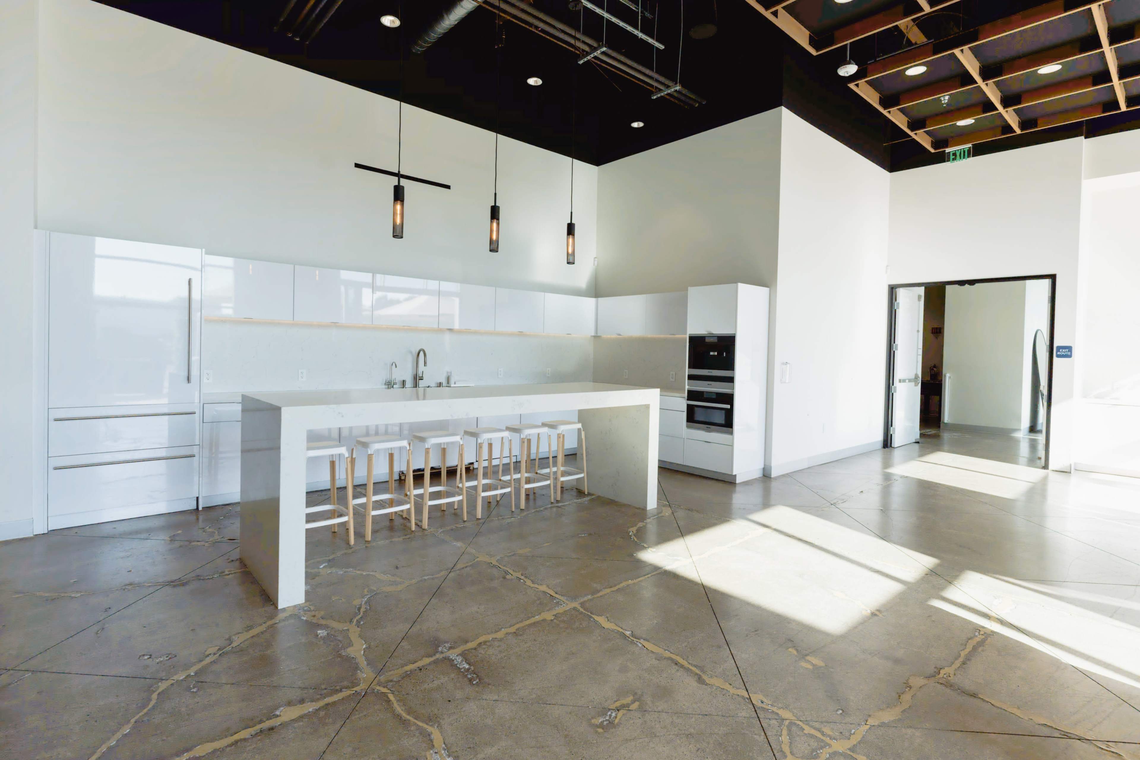 The image shows a modern kitchen with a large island, white cabinetry, and pendant lighting, situated in a spacious, well-lit room with polished concrete floors.