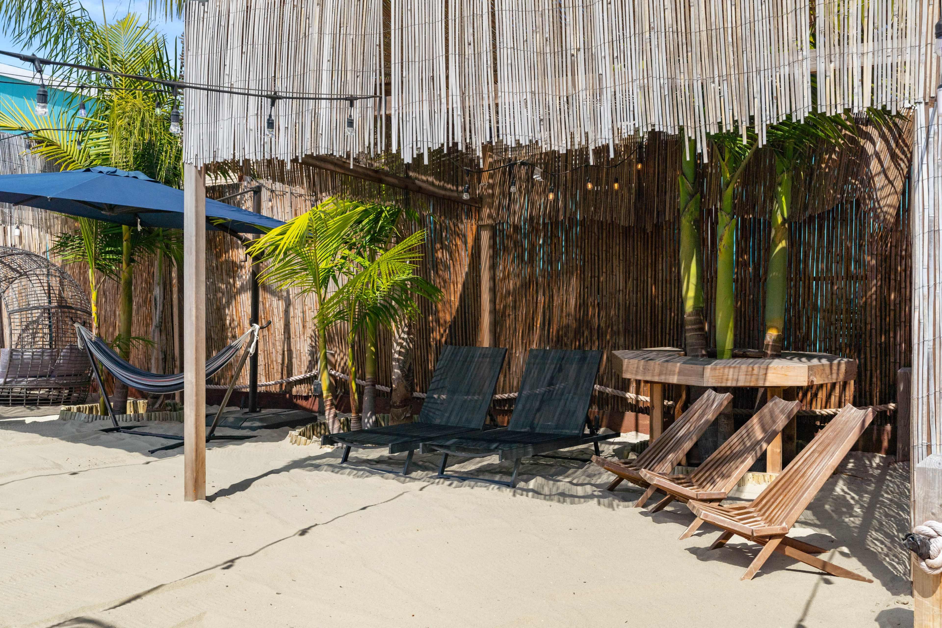 The image depicts a sandy beach area with lounge chairs, a hammock, a round wooden table, and palm trees under a thatched roof.