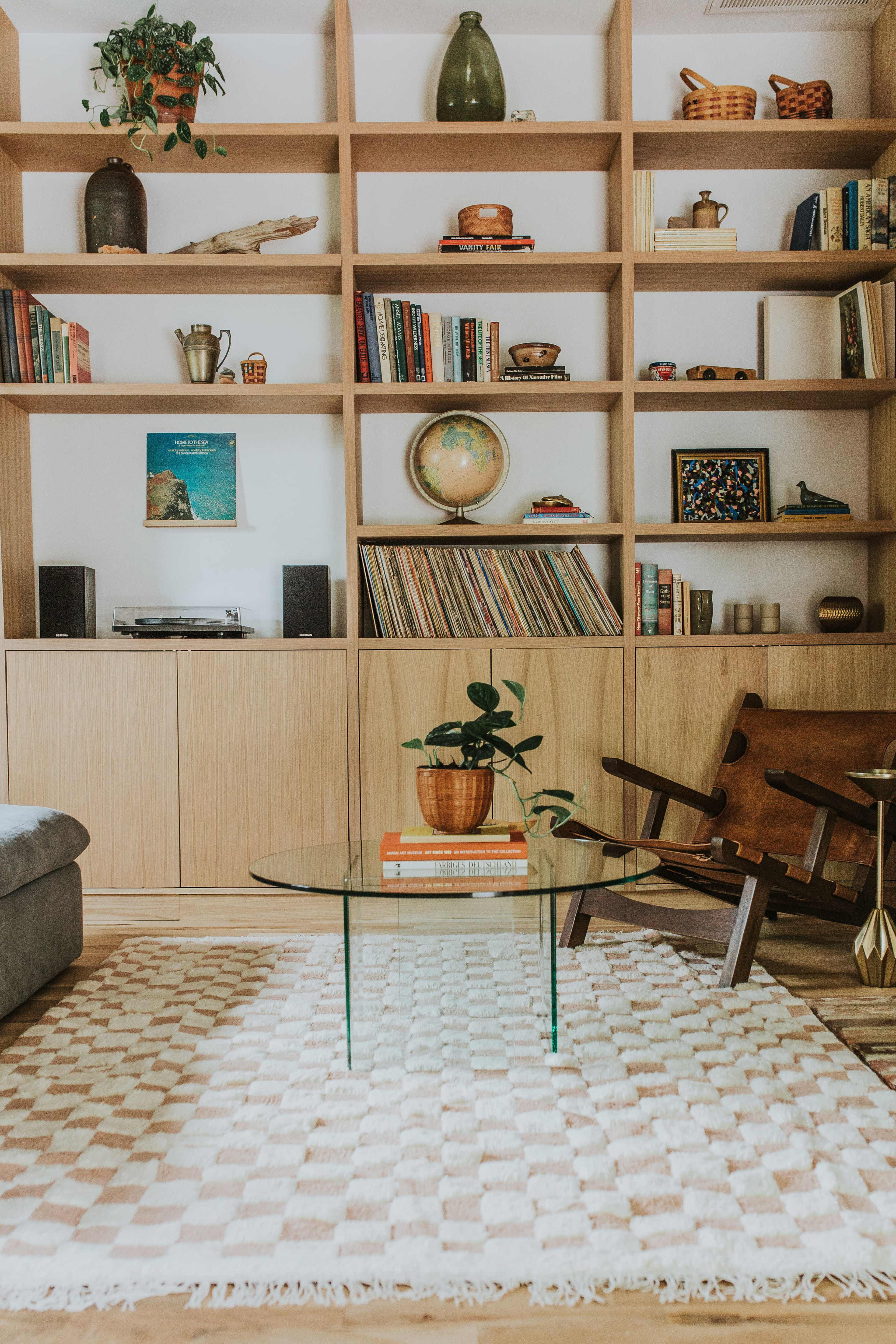 A living room with a wooden bookshelf filled with books and decorative items, a glass coffee table in the center, a patterned rug, and a comfortable seating area.