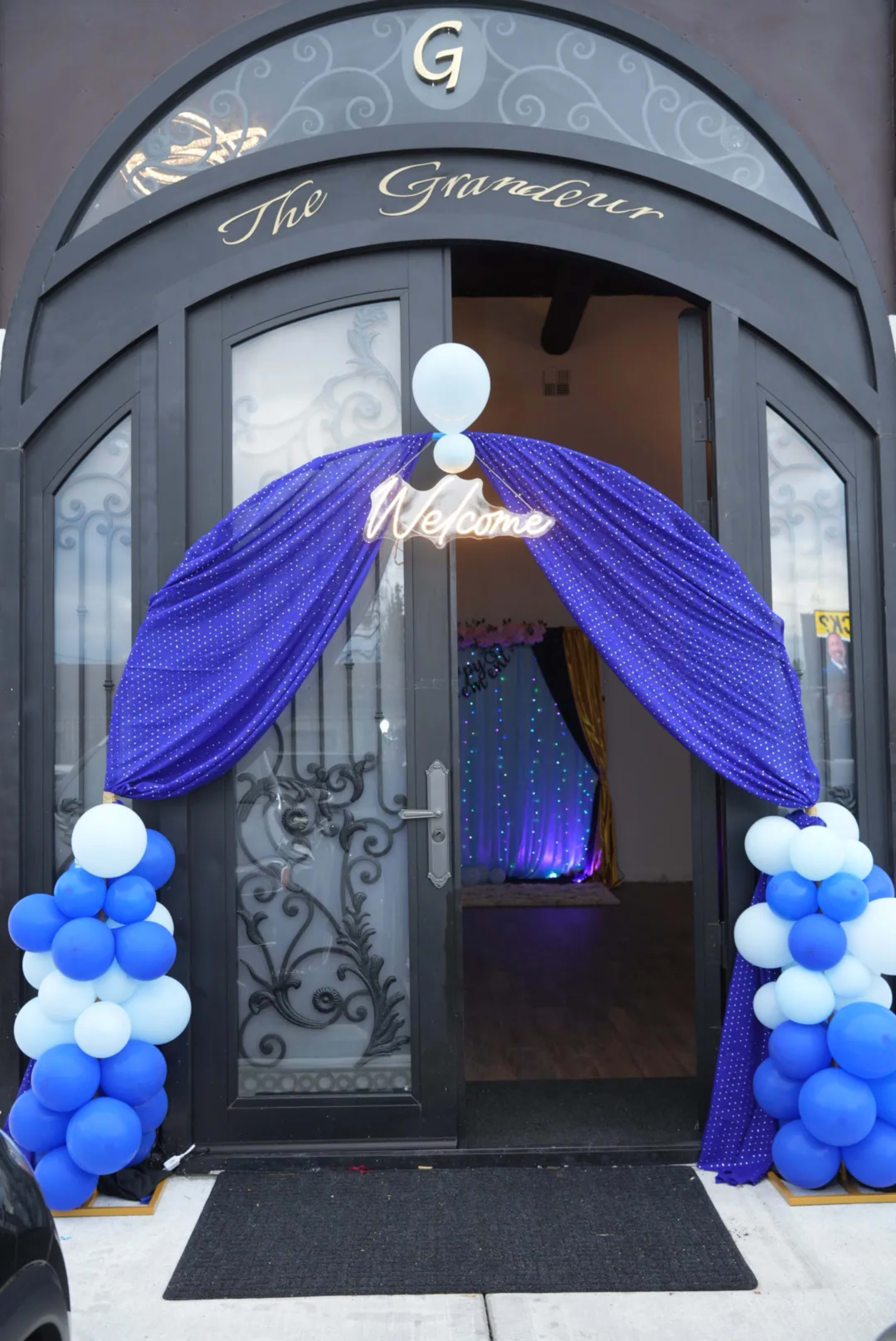 The entrance features a dark double door draped with blue and white balloons, welcoming guests with a "Welcome" sign illuminated above.