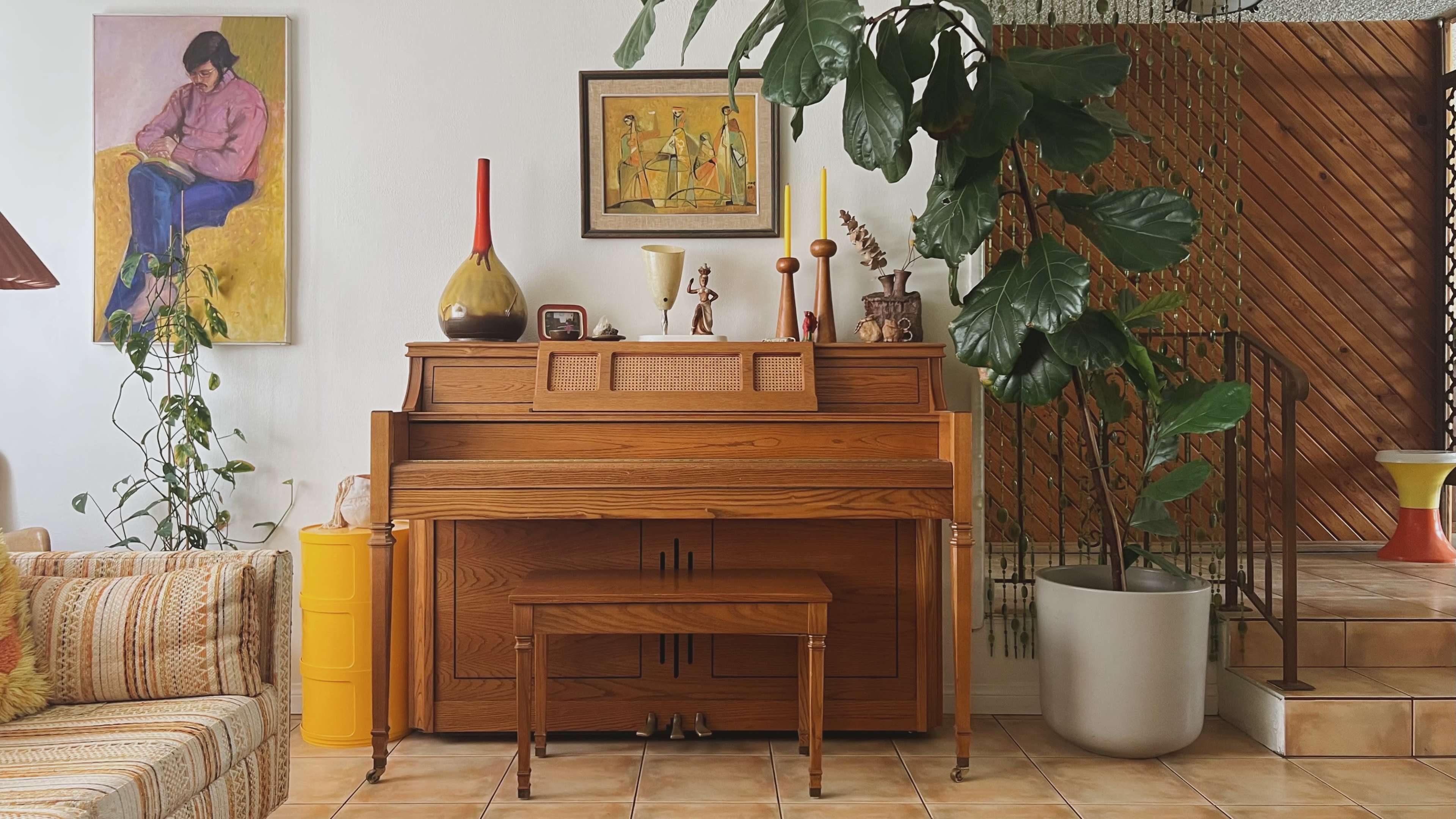 A wooden piano with a matching bench, surrounded by plants and artwork on the walls in a cozy living room setting.