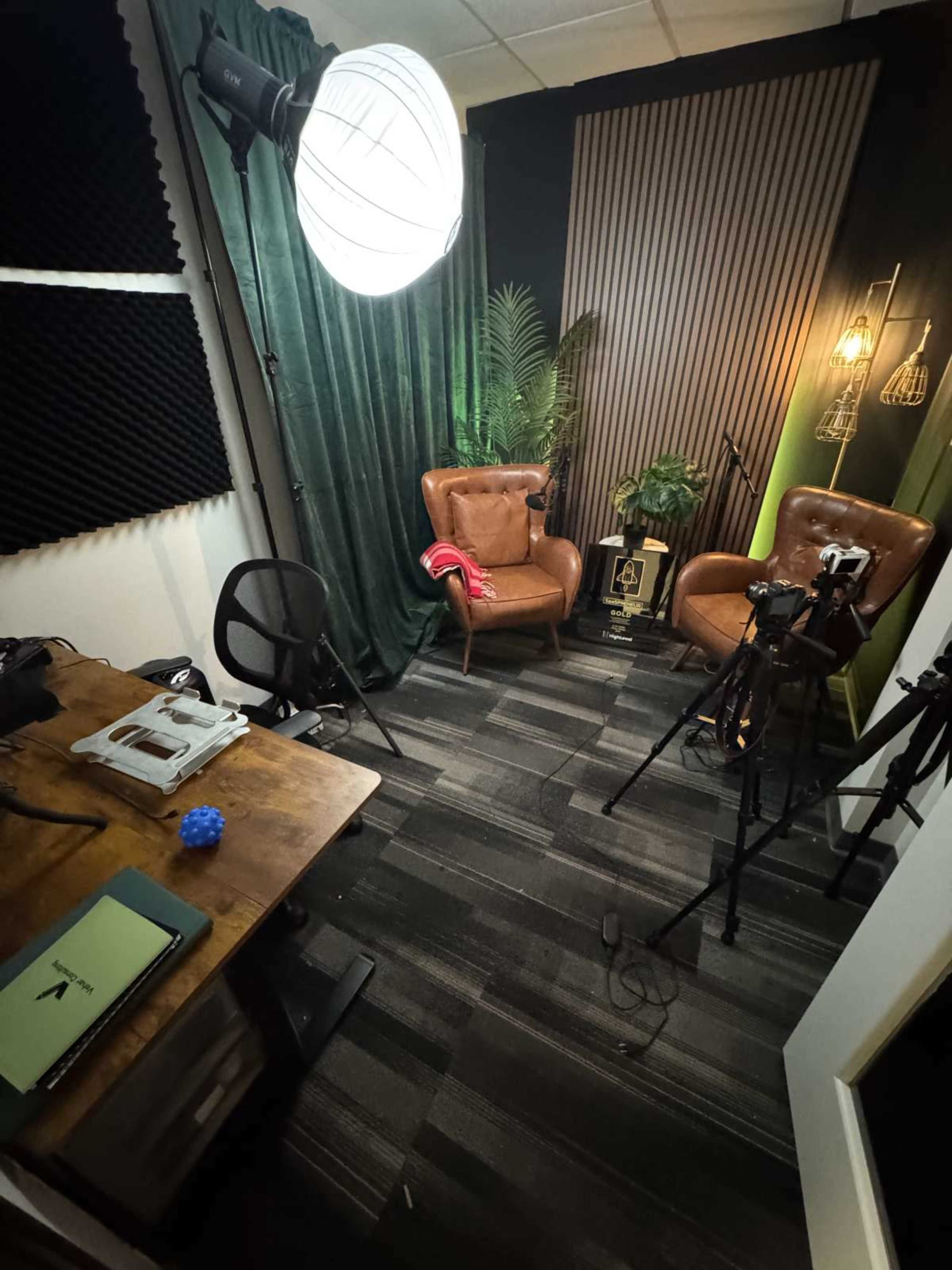 The image shows a small, well-lit studio space featuring two brown leather chairs, a green curtain, a wooden table, and several cameras and lighting equipment set up for video recording.