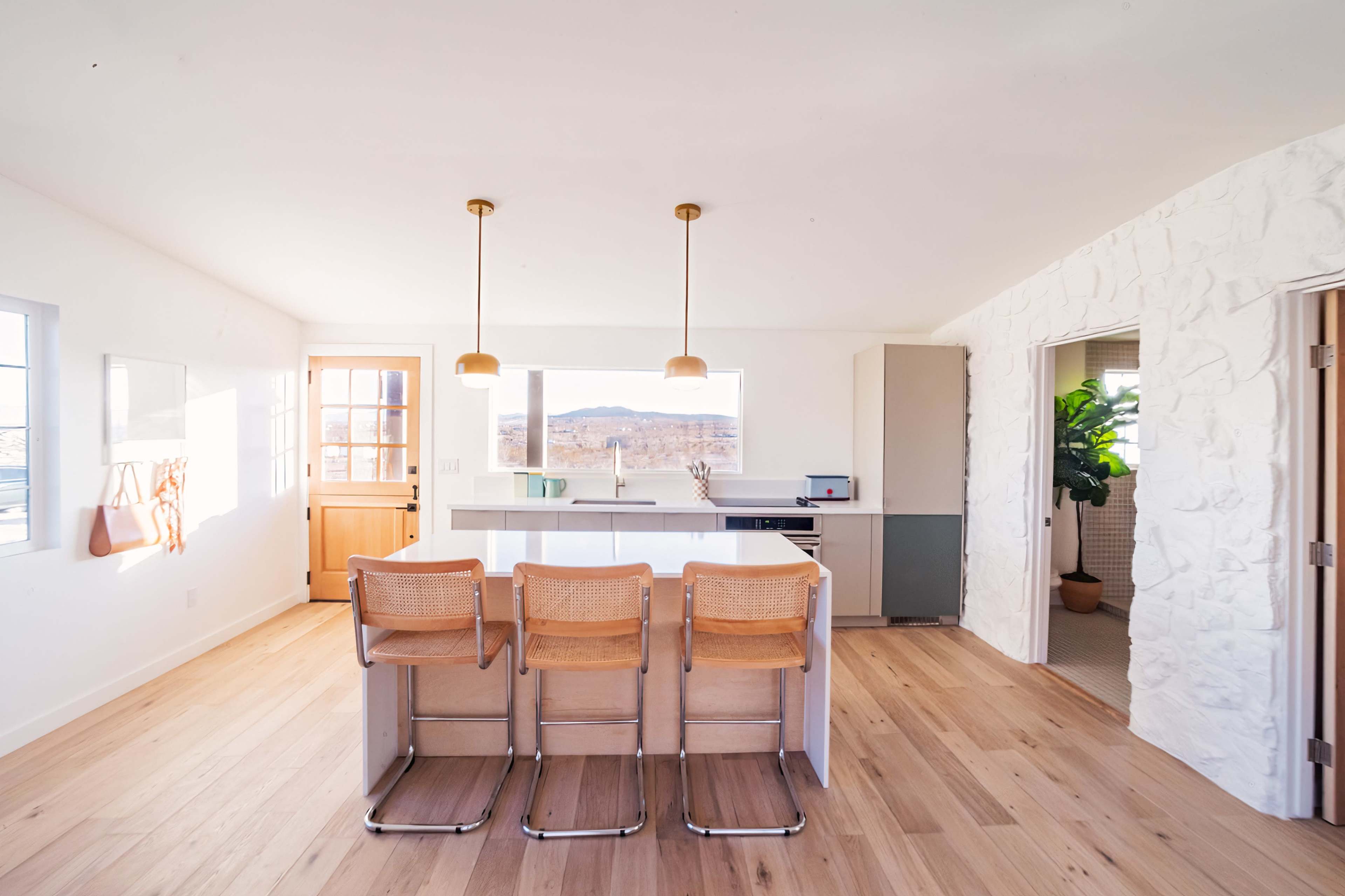 A modern kitchen with a light color scheme, featuring a central island with three bar stools, large windows, and a door leading outside.
