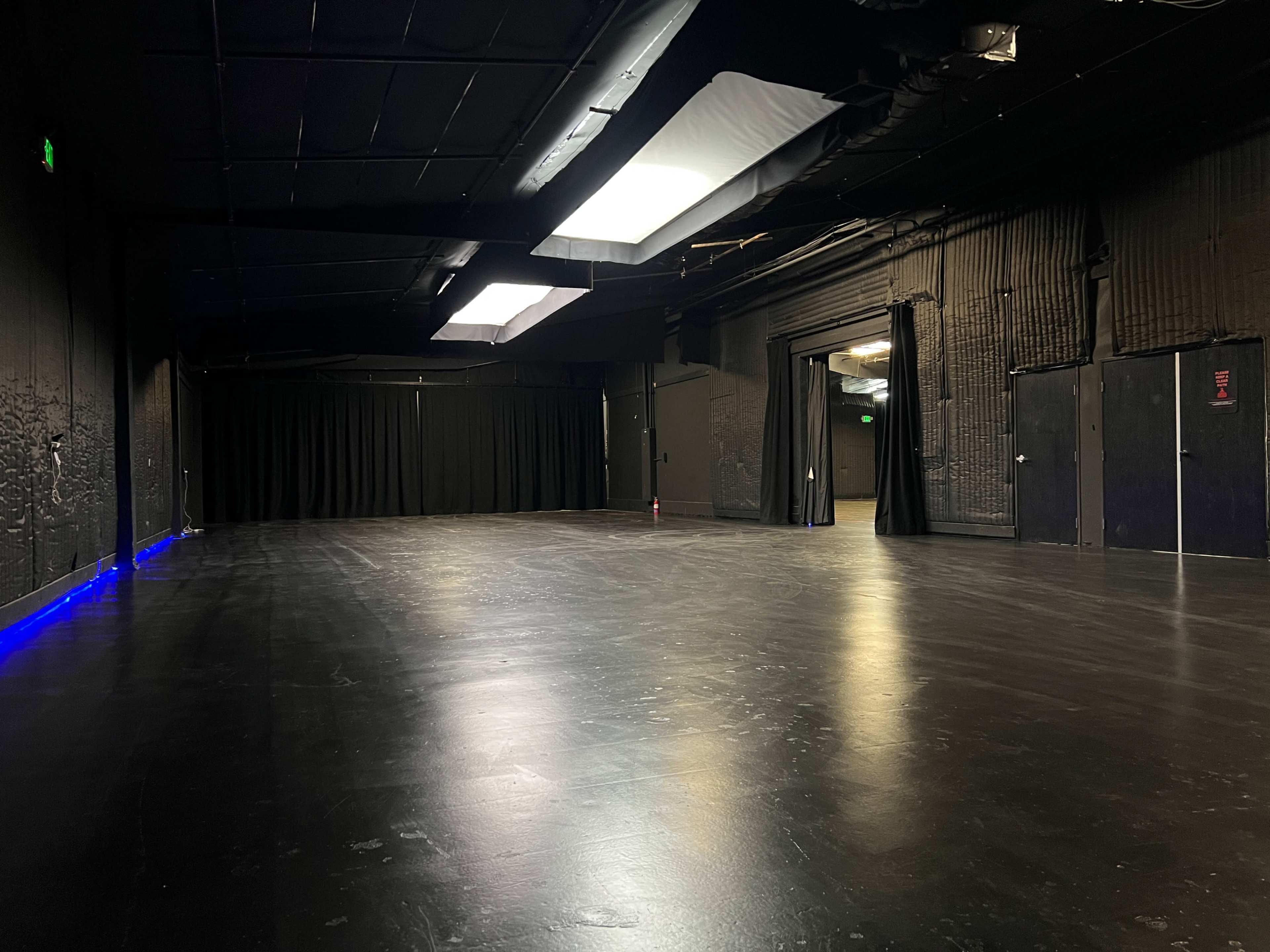 The image shows an empty black box theater with dark walls, a polished floor, and dim lighting.