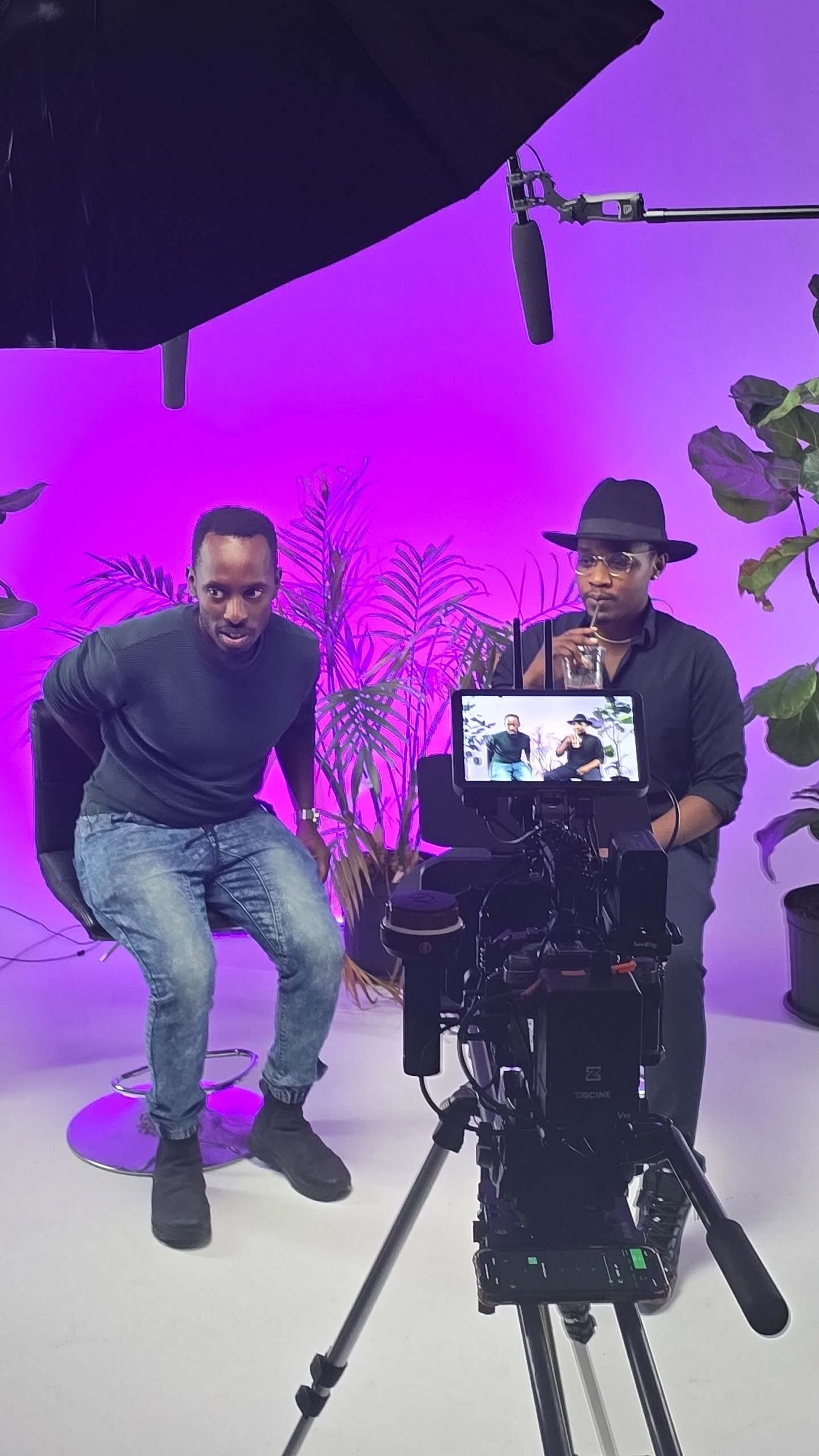Two men are seated in a studio setting surrounded by plants, with a camera focused on them and colorful lighting in the background.