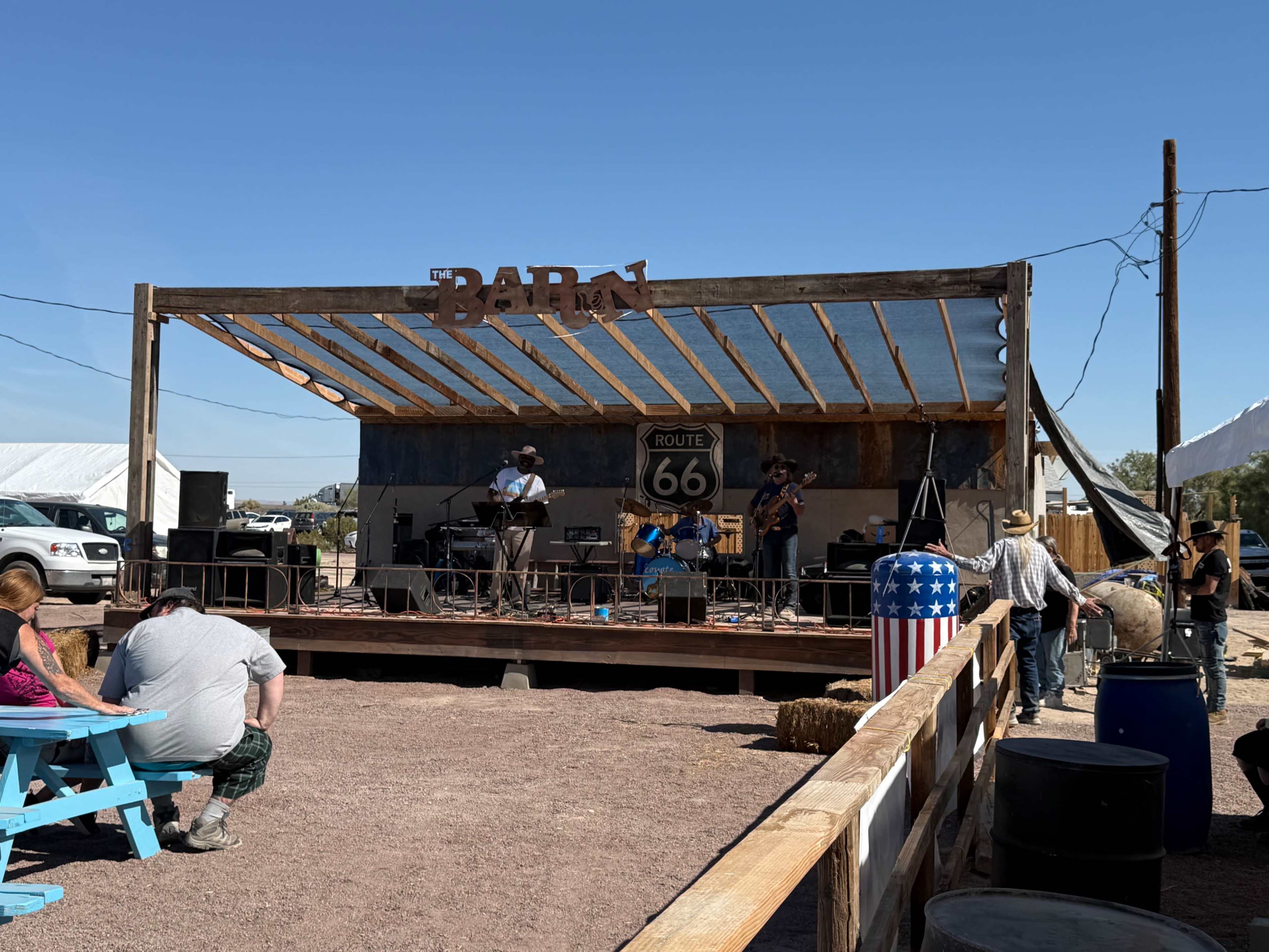 A band performs on a wooden stage at a Route 66 venue, while visitors gather around in a desert setting.