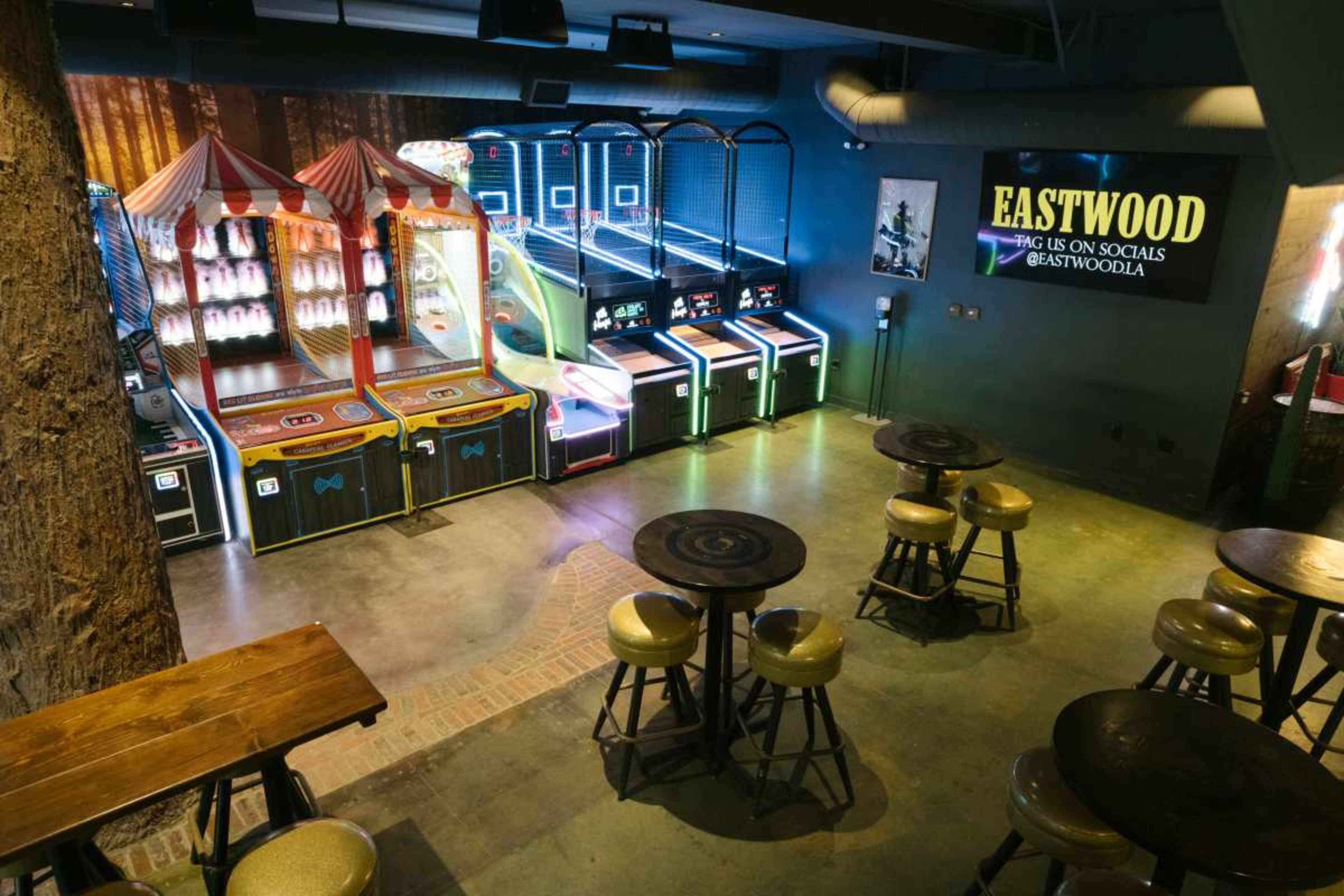 The image shows an entertainment area featuring arcade games, a circus-themed game booth, and several tables with chairs arranged in a dimly lit space.
