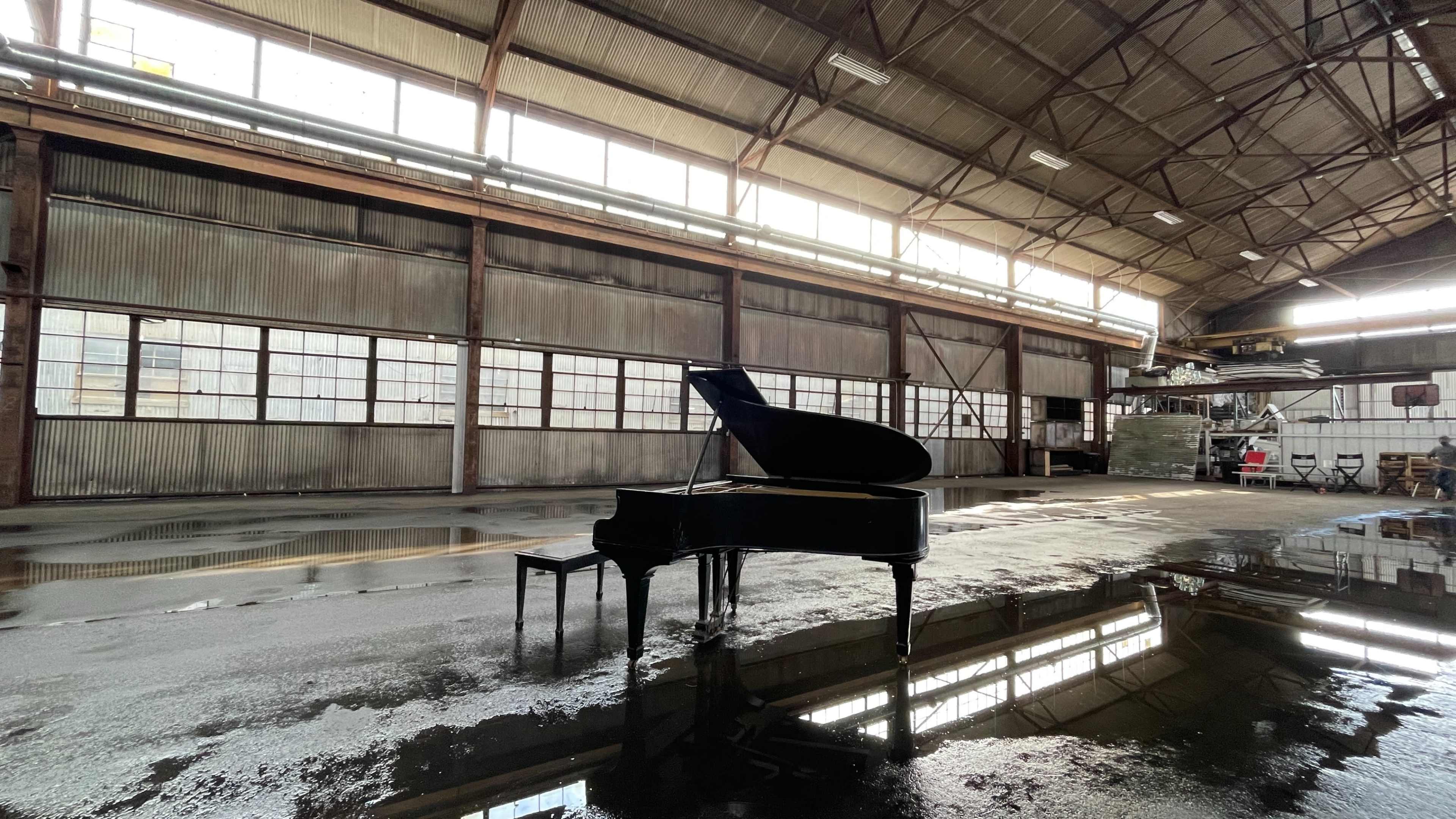 A black grand piano is positioned on a wet concrete floor in a large, industrial warehouse with high ceilings and numerous windows.