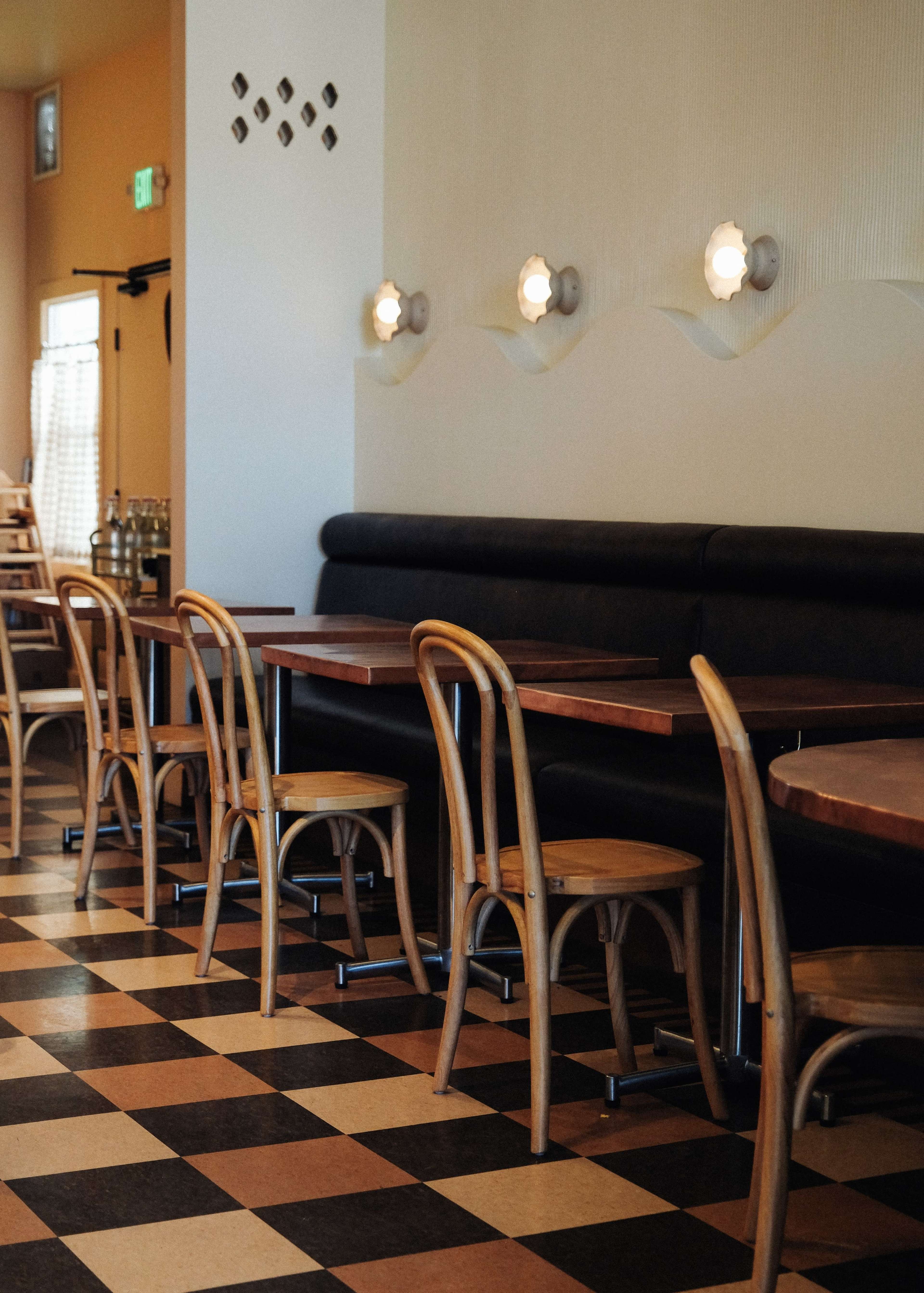A sparsely furnished restaurant features wooden tables and bentwood chairs arranged on a checkered floor.