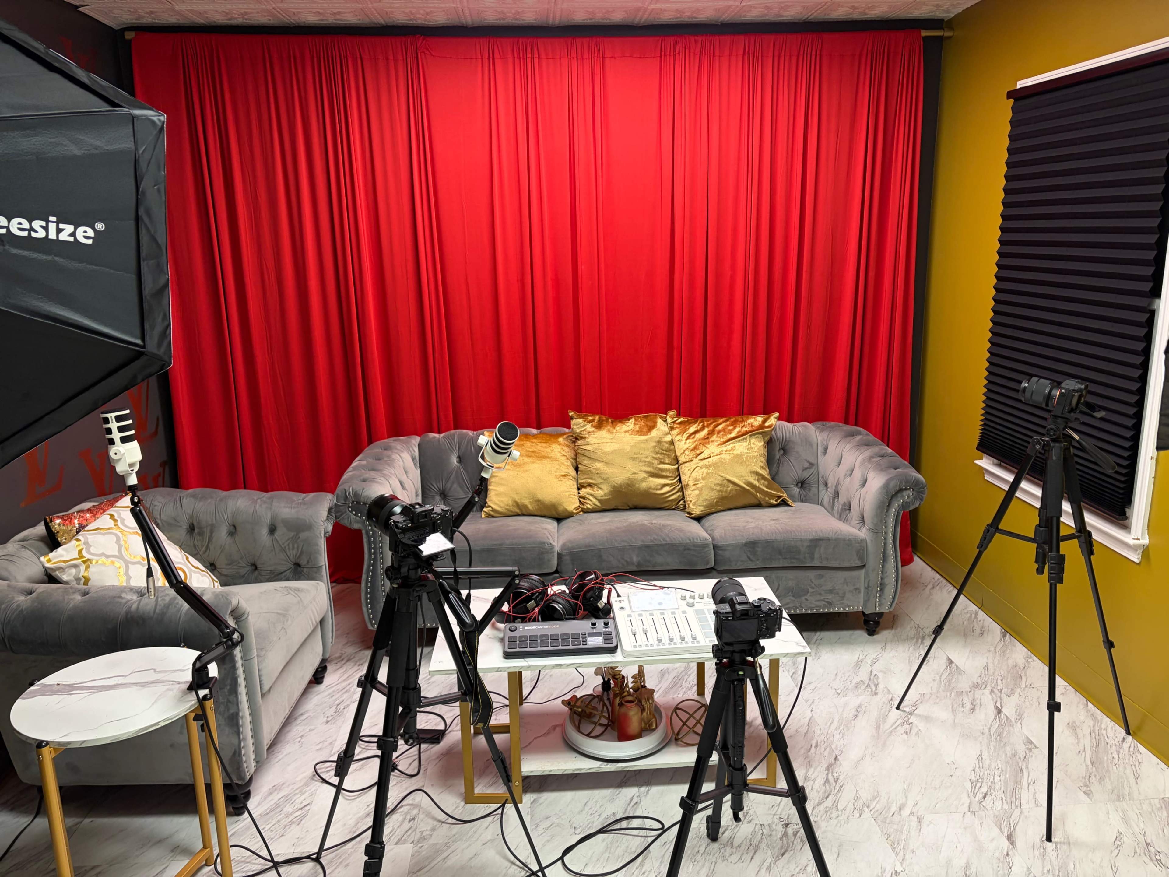 The image shows a modern studio setup with a gray sofa, red curtains, and cameras positioned on tripods around a central table with equipment.