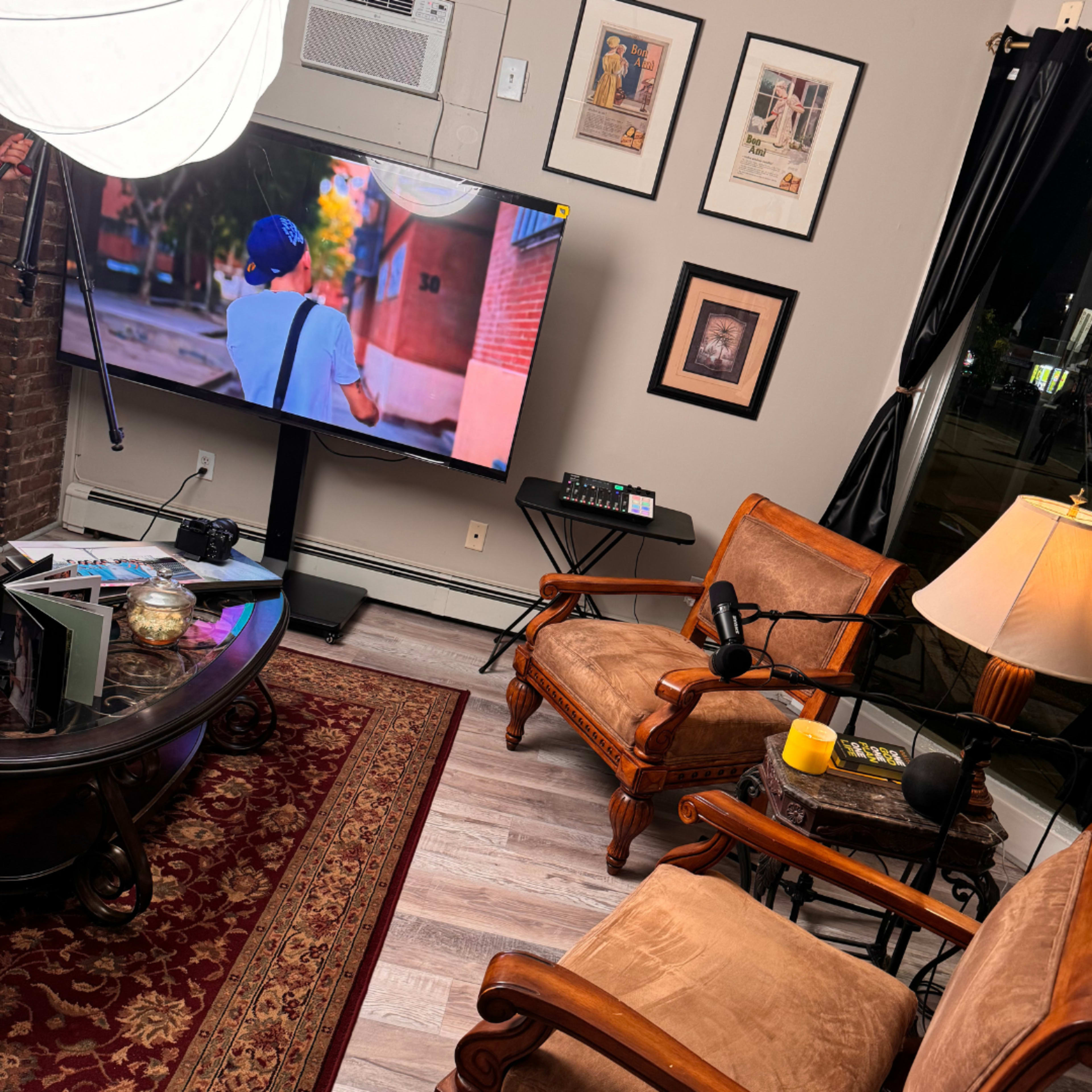 The image shows a cozy living room setting with two wooden chairs facing a television, a coffee table in the center, and decor including framed pictures on the walls.