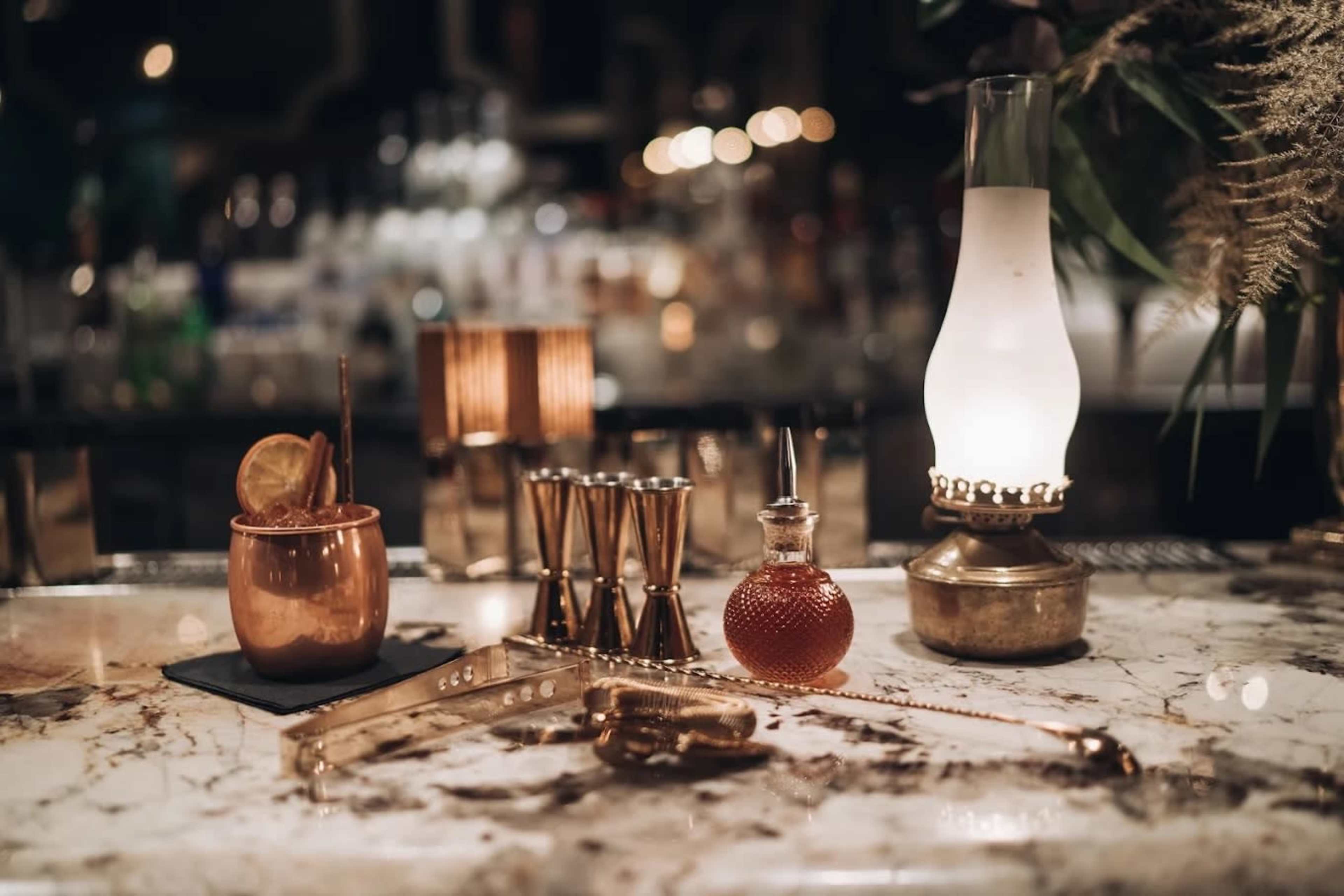 The image showcases a sophisticated bar scene featuring a copper mug, jiggers, a decorative bottle, and an antique lamp on a marble countertop.