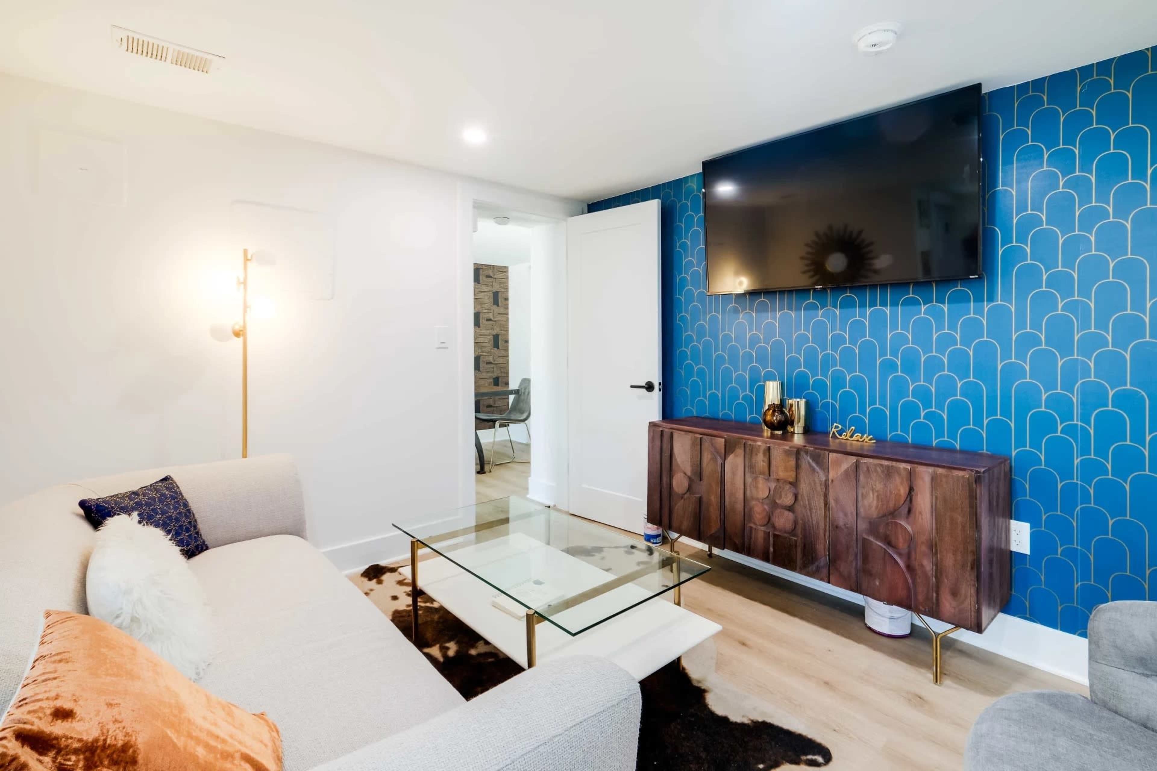 A modern living room with a light gray sofa, a glass coffee table, and a wall-mounted flat-screen TV above a wooden cabinet with decorative items.