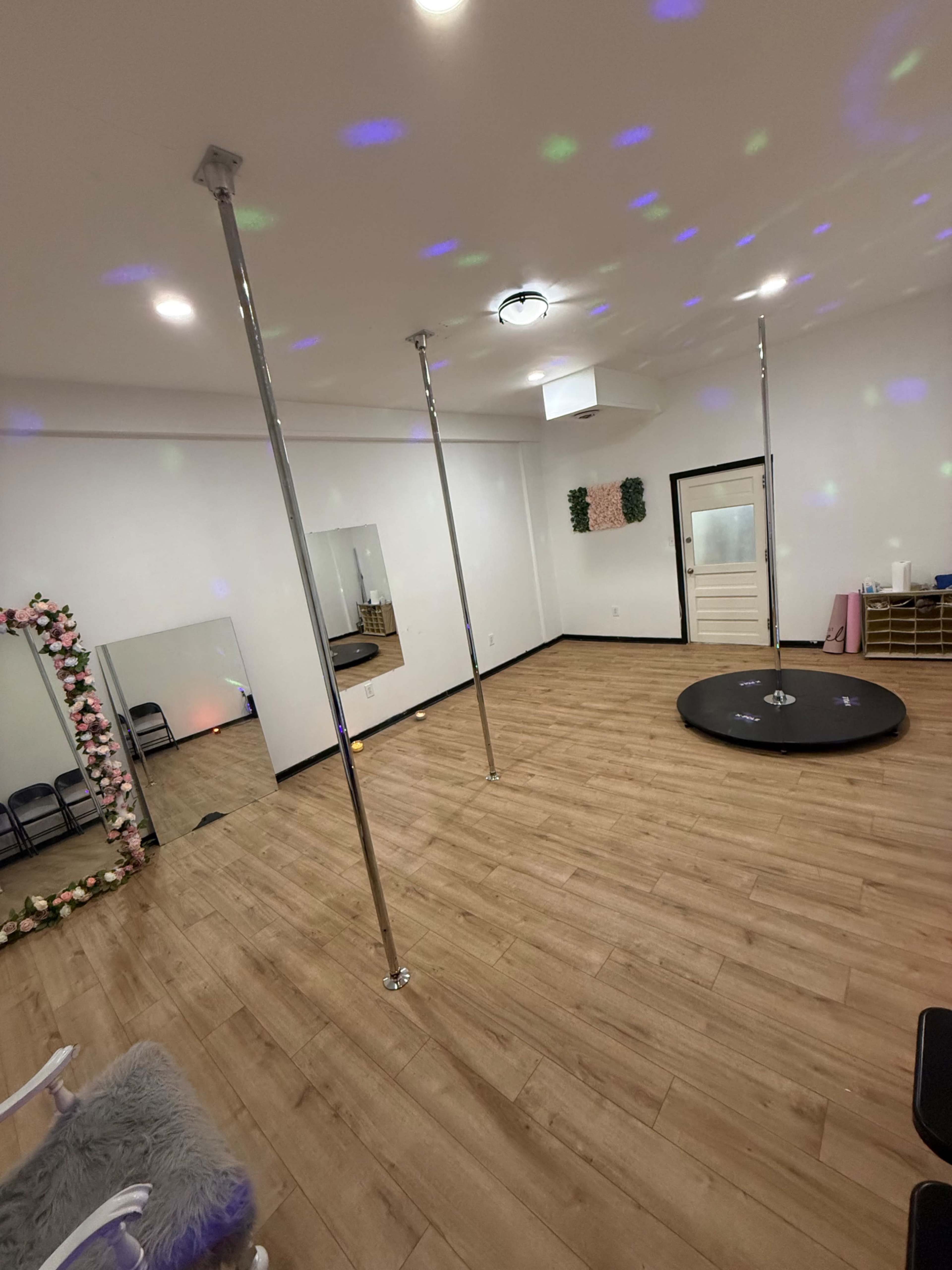 The image shows an empty dance studio featuring two mirrored poles, a round dance floor, and soft lighting effects on the walls.