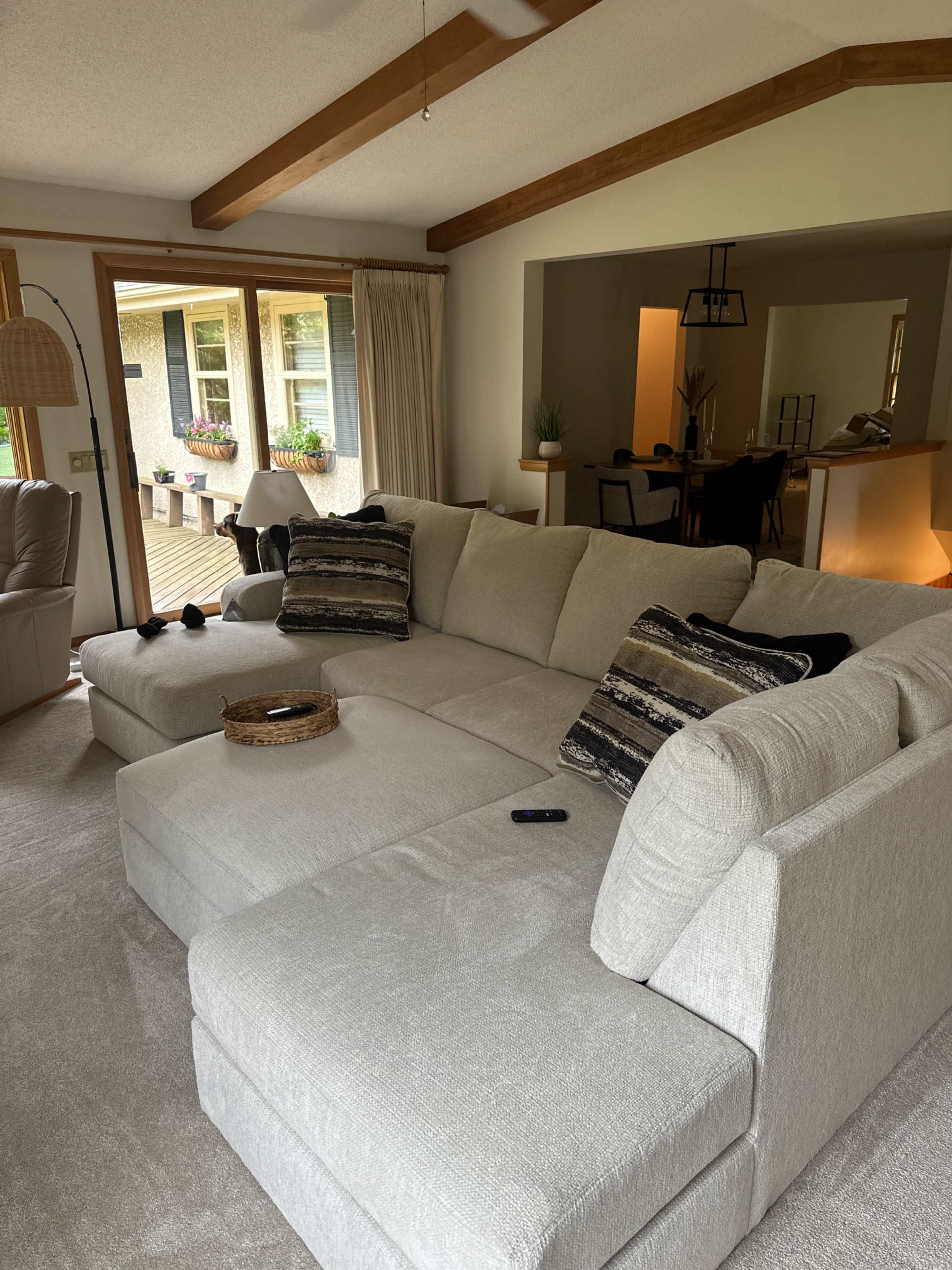 The image shows a light-colored sectional sofa in a living room with large windows and a dining area visible in the background.