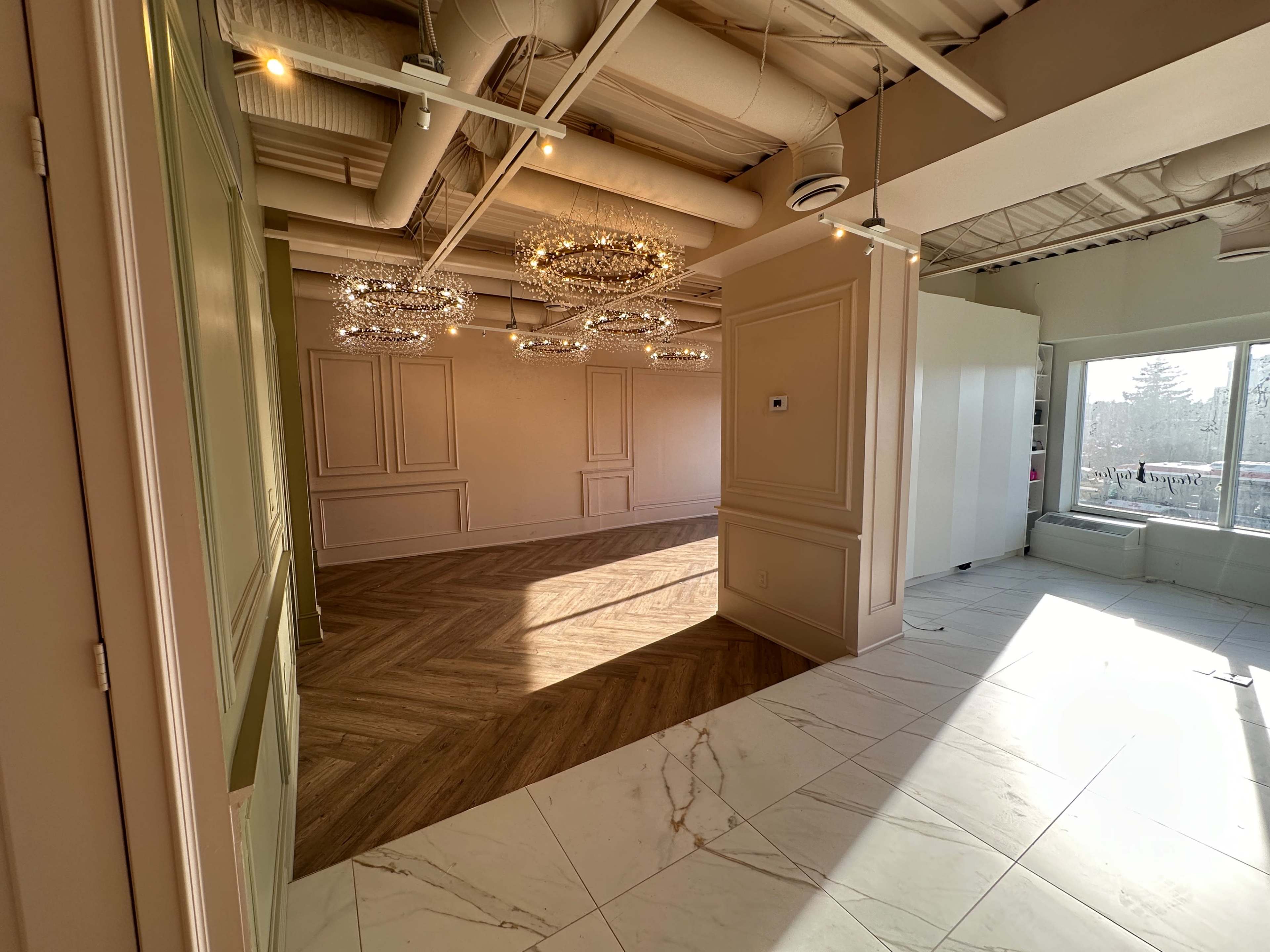 A spacious interior room featuring decorative chandeliers, patterned flooring, and large windows allowing natural light to enter.