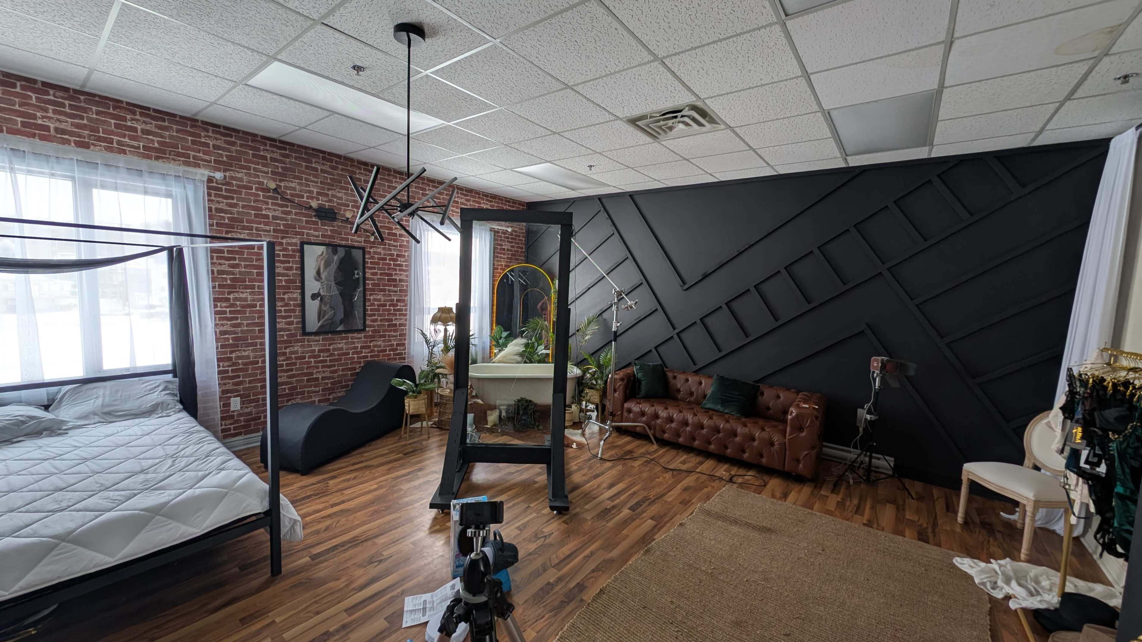 The image shows a spacious studio room featuring a four-poster bed, a dark accent wall with geometric patterns, a brown leather couch, and a large window allowing natural light.
