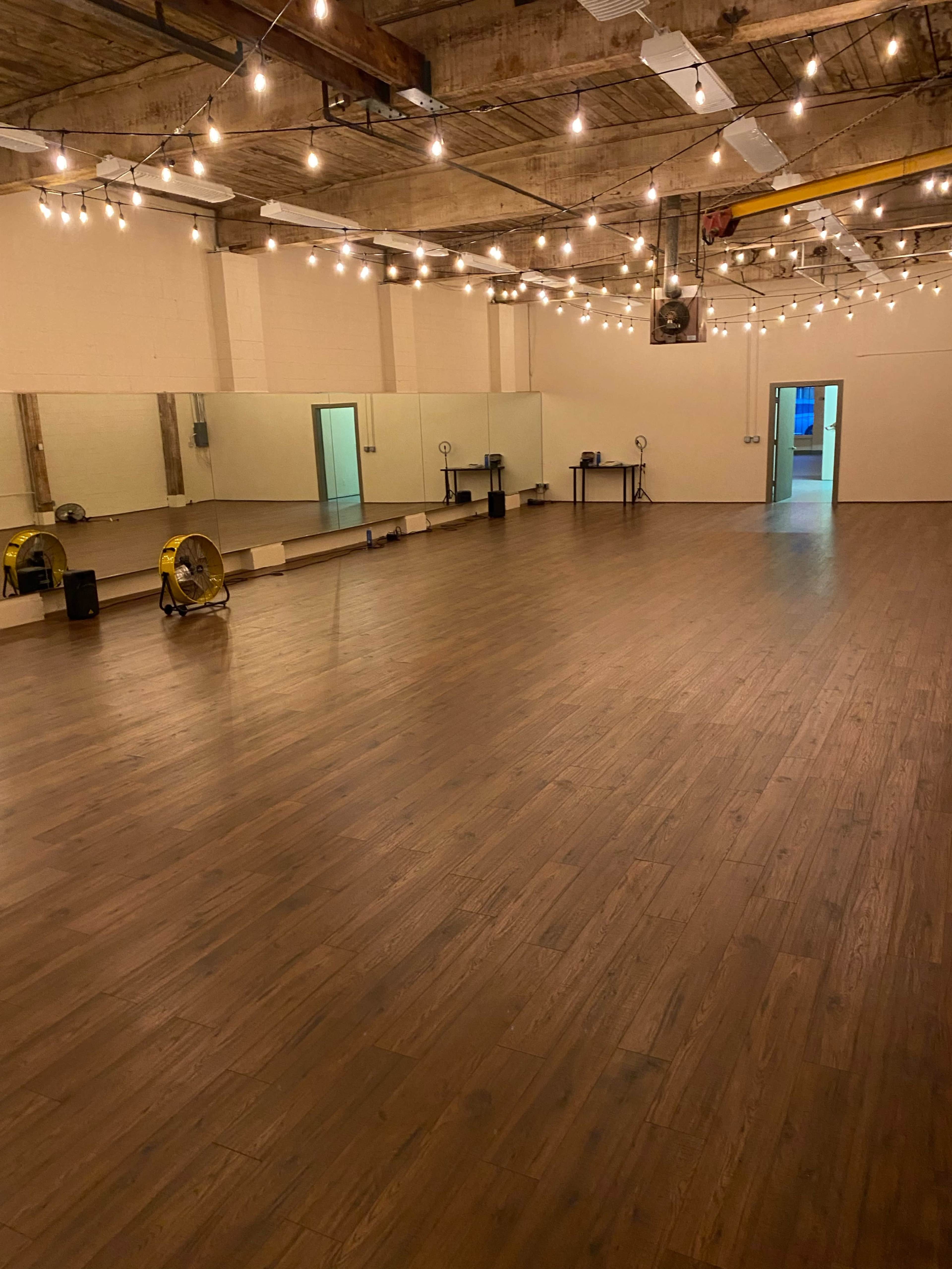 A spacious dance studio with wooden flooring, string lights overhead, and a mirrored wall.