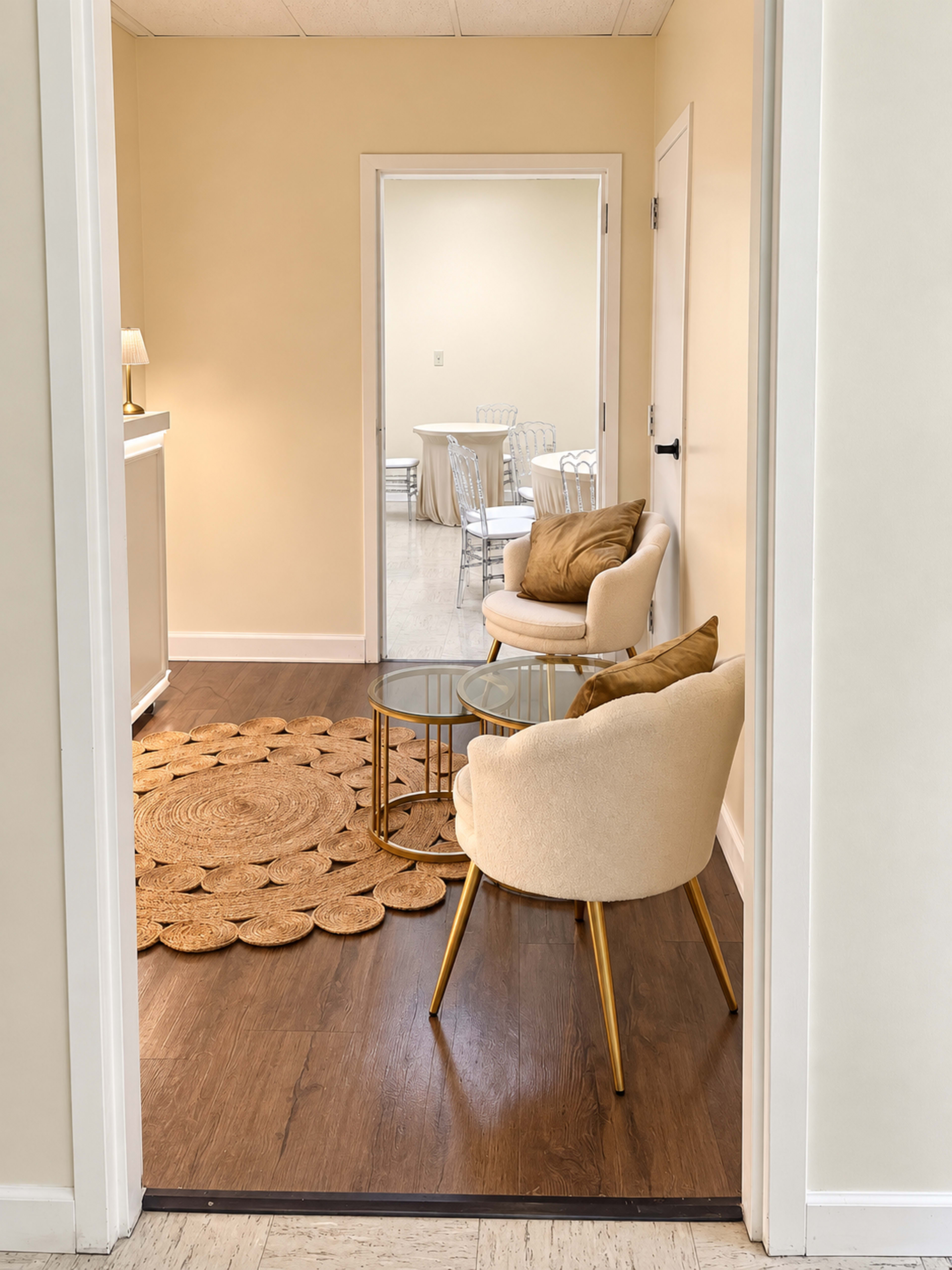 A small waiting area features two chairs and a round glass table, with a decorative rug on the floor and a view into an adjoining room with tables set up.