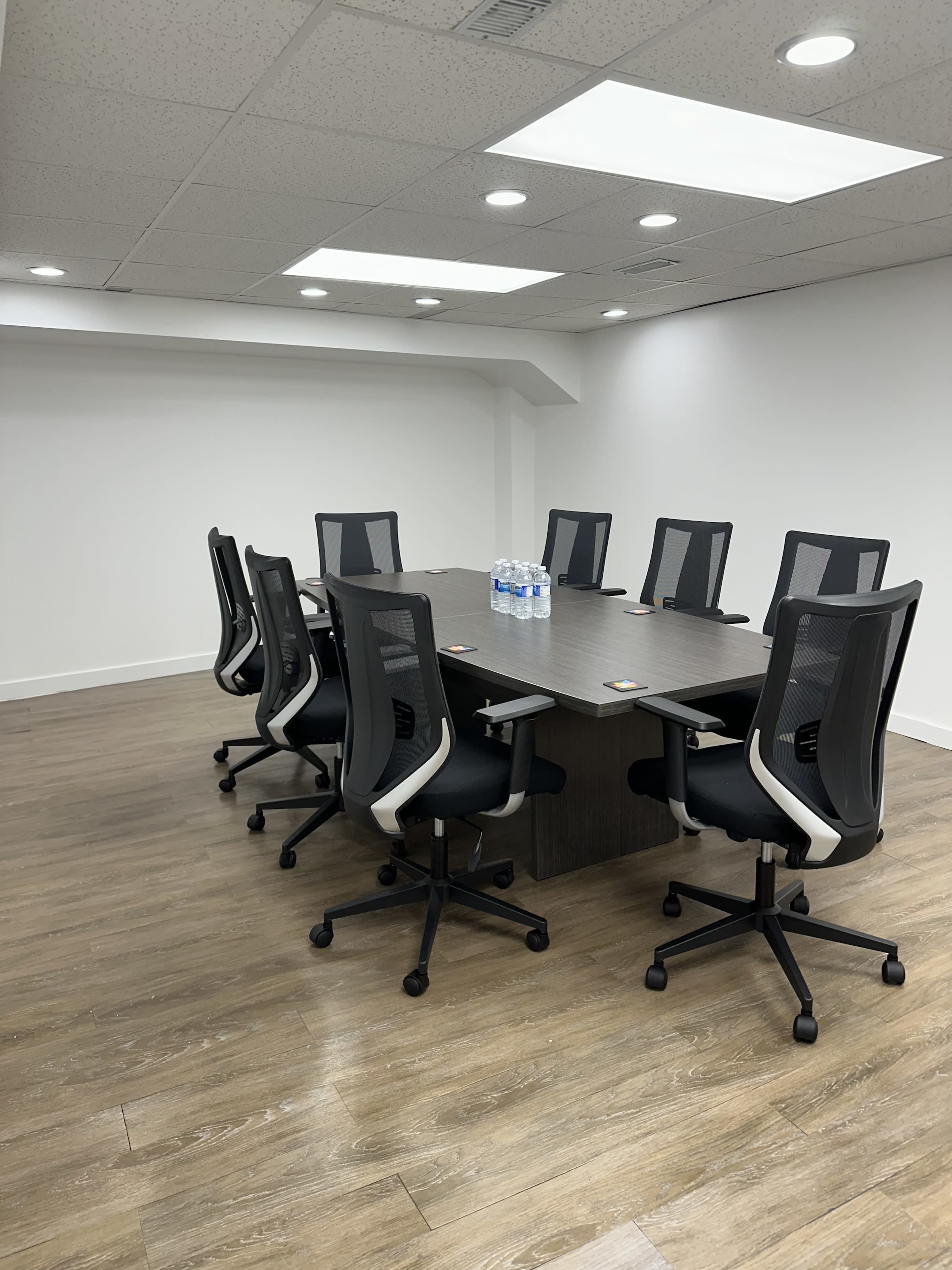 A conference room features a long table surrounded by eight ergonomic chairs and a few bottles of water on the table.