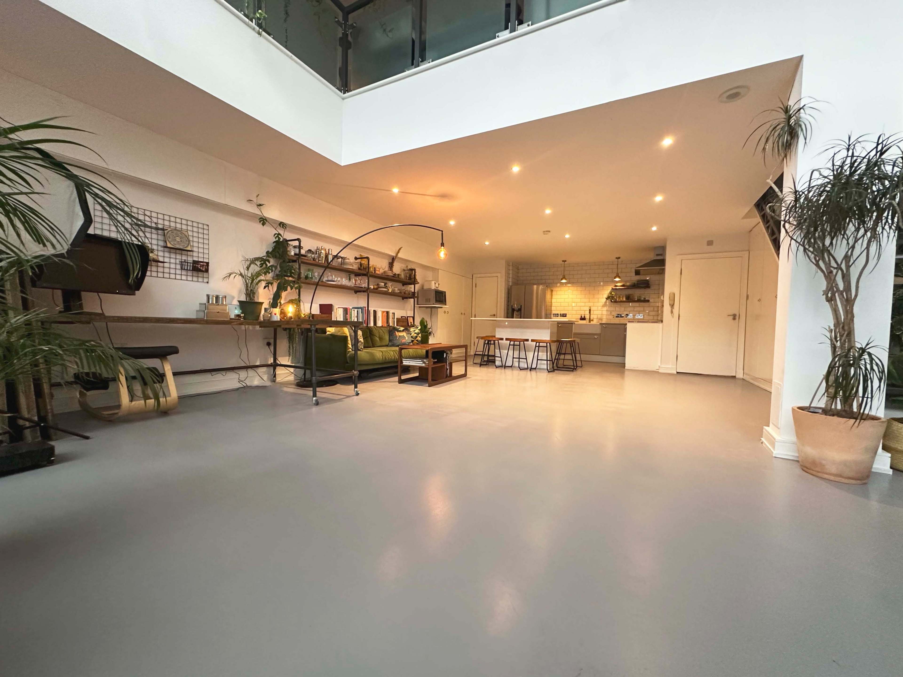 A spacious, modern room featuring a green sofa, a wooden table with stools, and a well-lit kitchen area in the background.