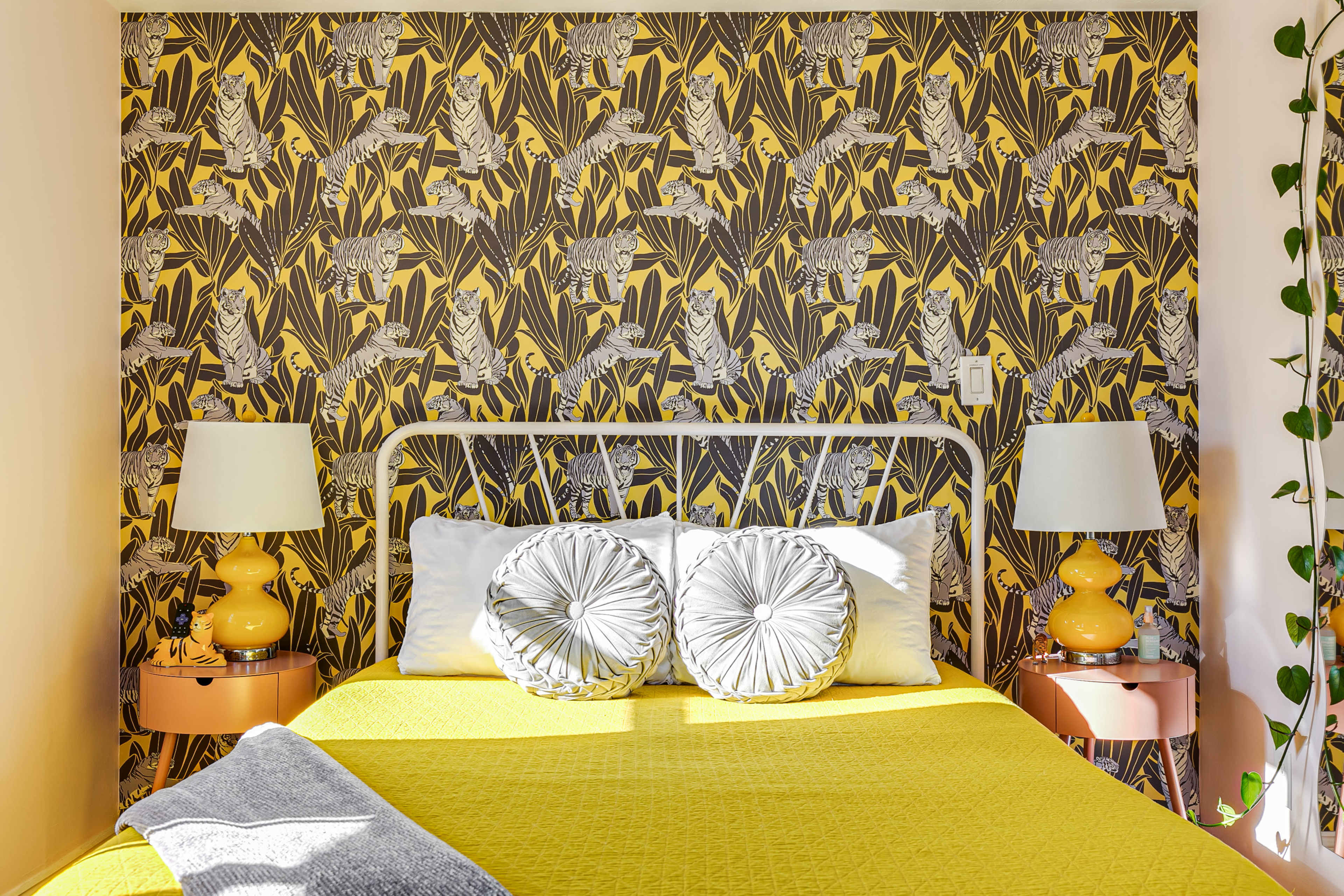 A bright yellow bedspread with two decorative pillows is positioned against a patterned wallpaper featuring tropical leaves and birds, flanked by matching lamps on side tables.