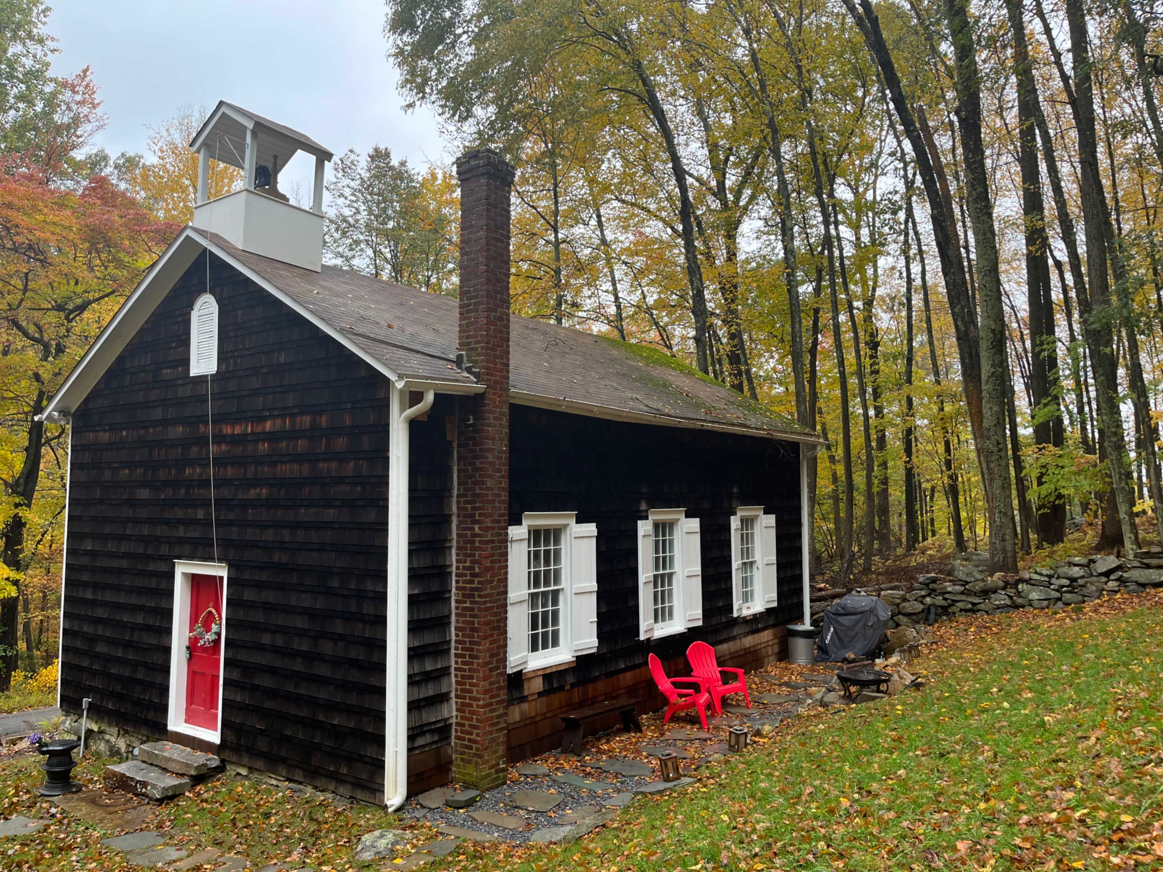 A rustic cabin with a red door and two red chairs sits beneath a canopy of autumn trees.