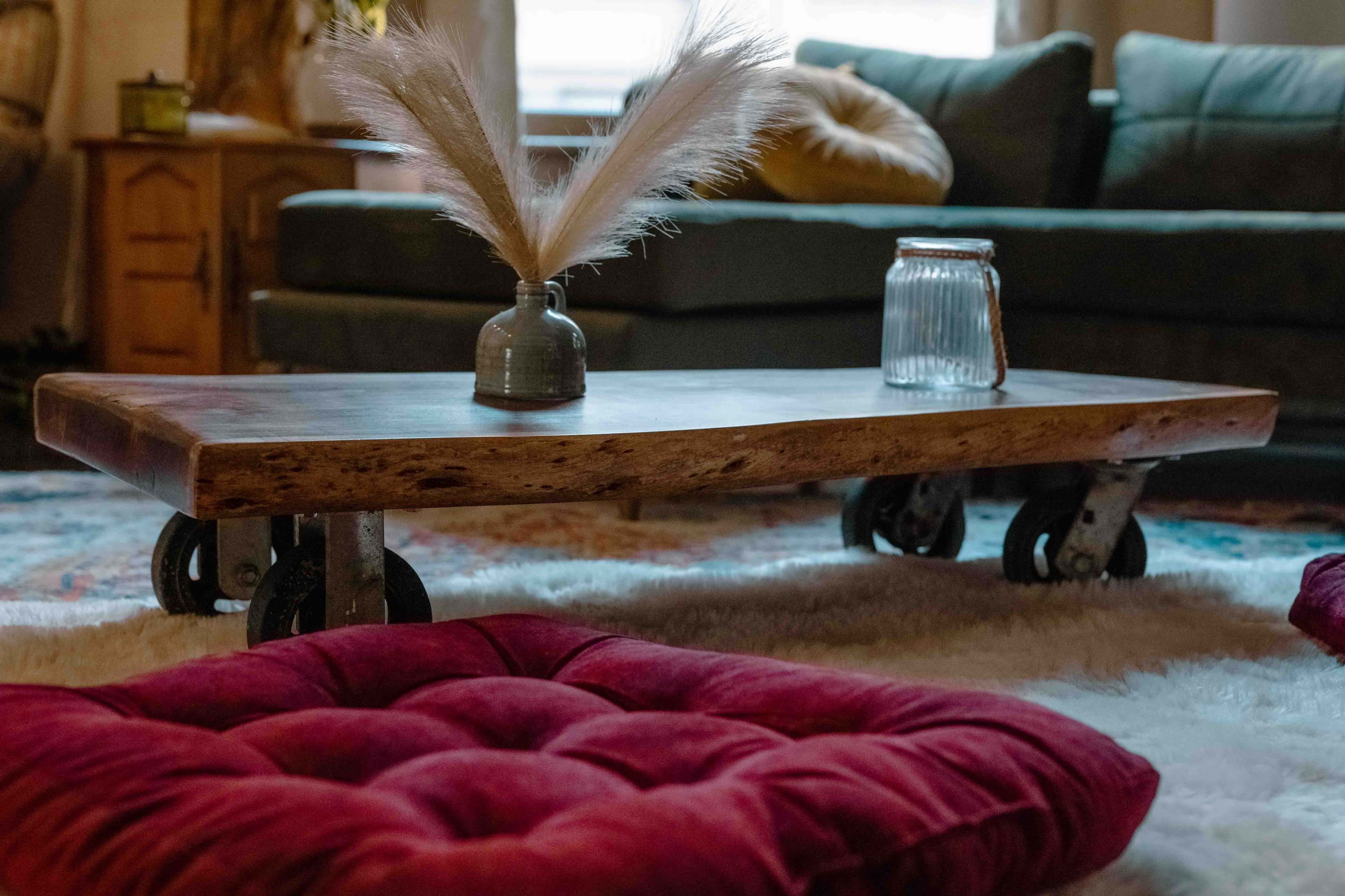 A wooden coffee table on wheels is positioned in a living room, with a small vase and a glass jar atop it, surrounded by plush, red cushions on a plush rug.