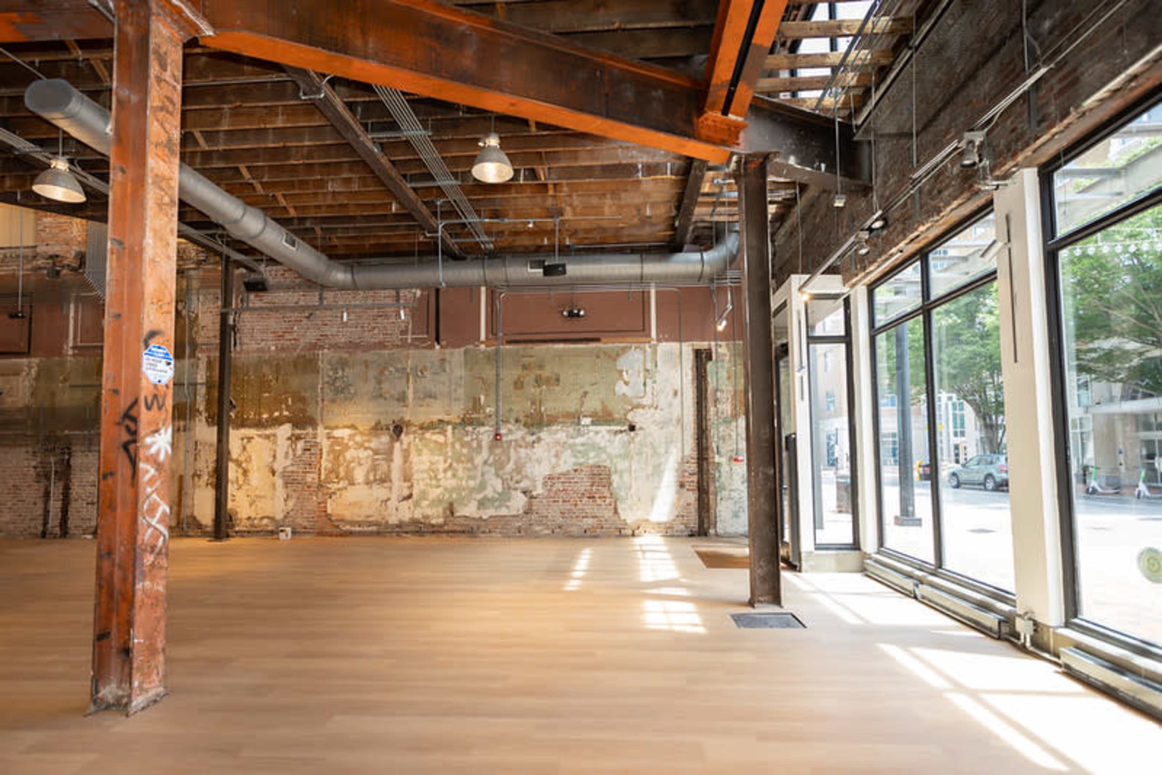 The image shows a spacious, industrial-style interior with exposed brick walls and wooden beams, featuring large windows that allow natural light to illuminate the floor.