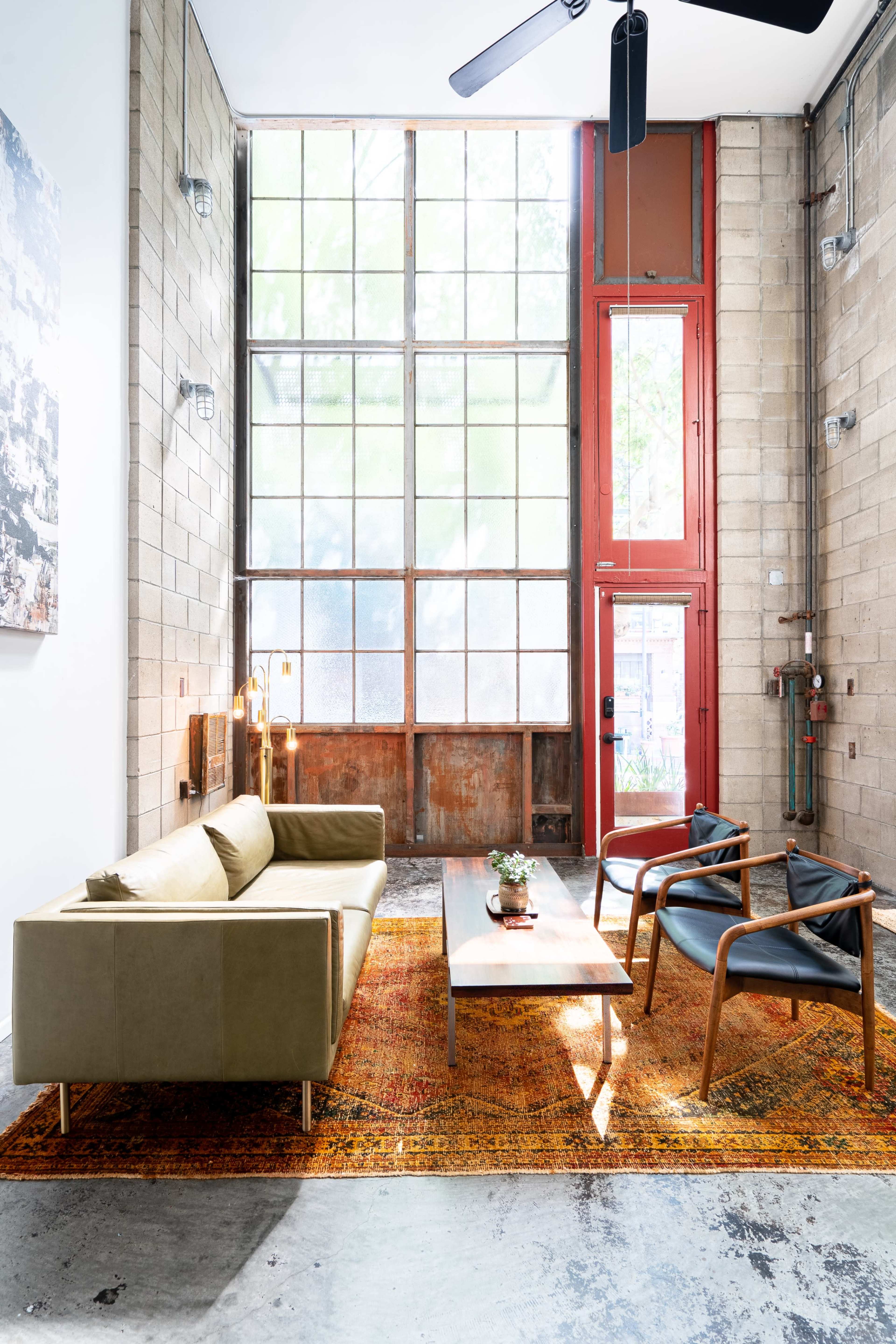 The image features a modern living room with a large window, a green sofa, two black chairs, a coffee table, and a colorful rug on a concrete floor.