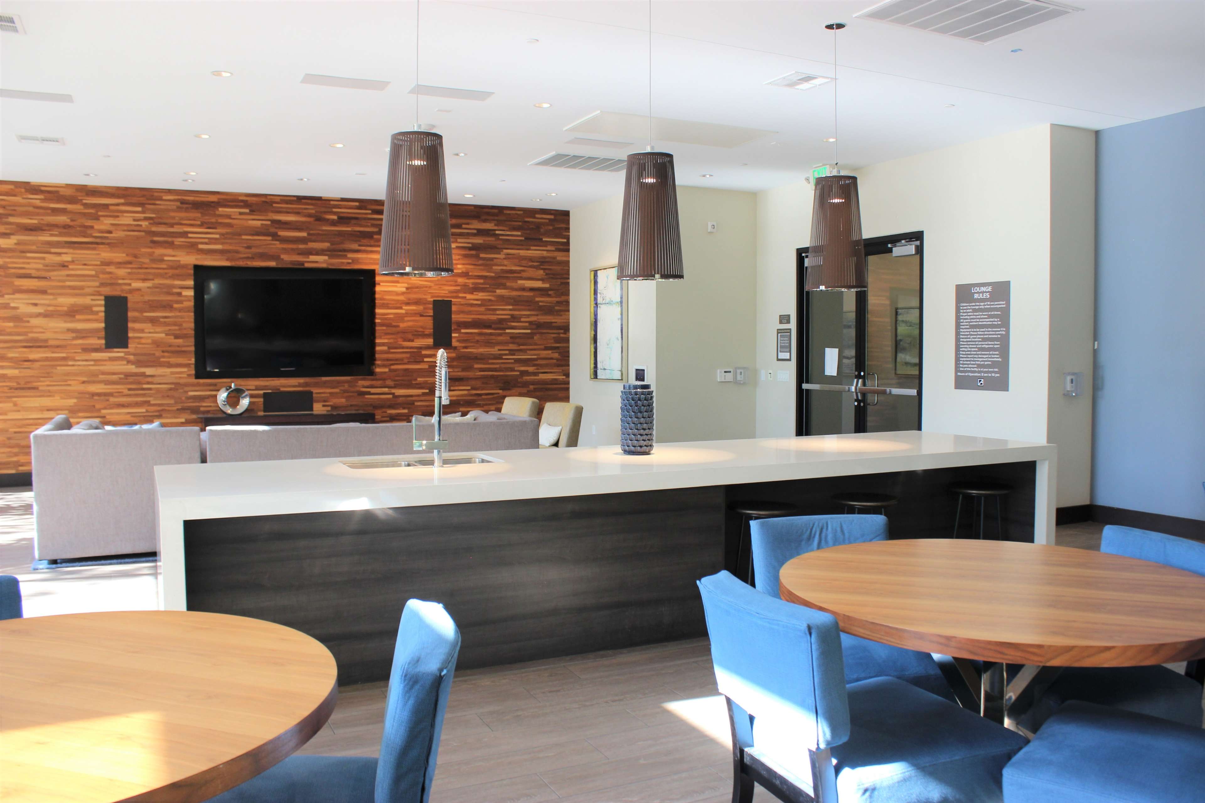 A modern communal lounge area featuring a kitchenette, tables with blue chairs, and a seating arrangement with a television on the wall.