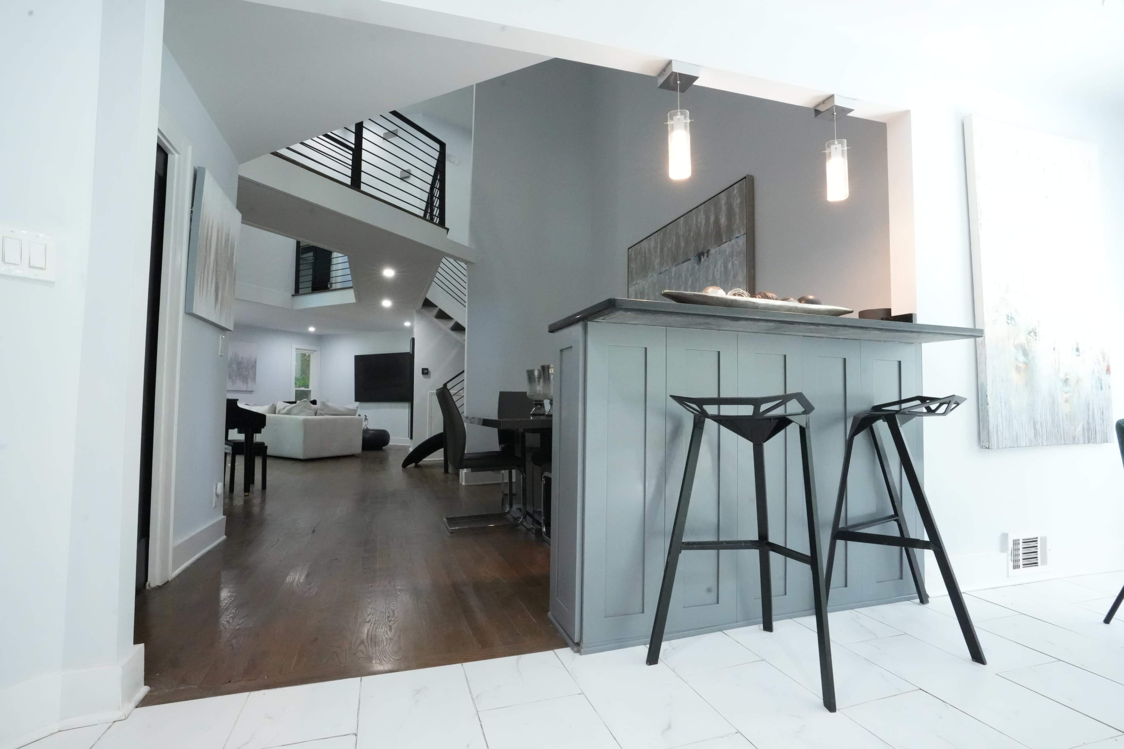 The image shows a modern interior space featuring a kitchen bar with two black stools, leading to an open floor plan that includes a staircase and a living area.