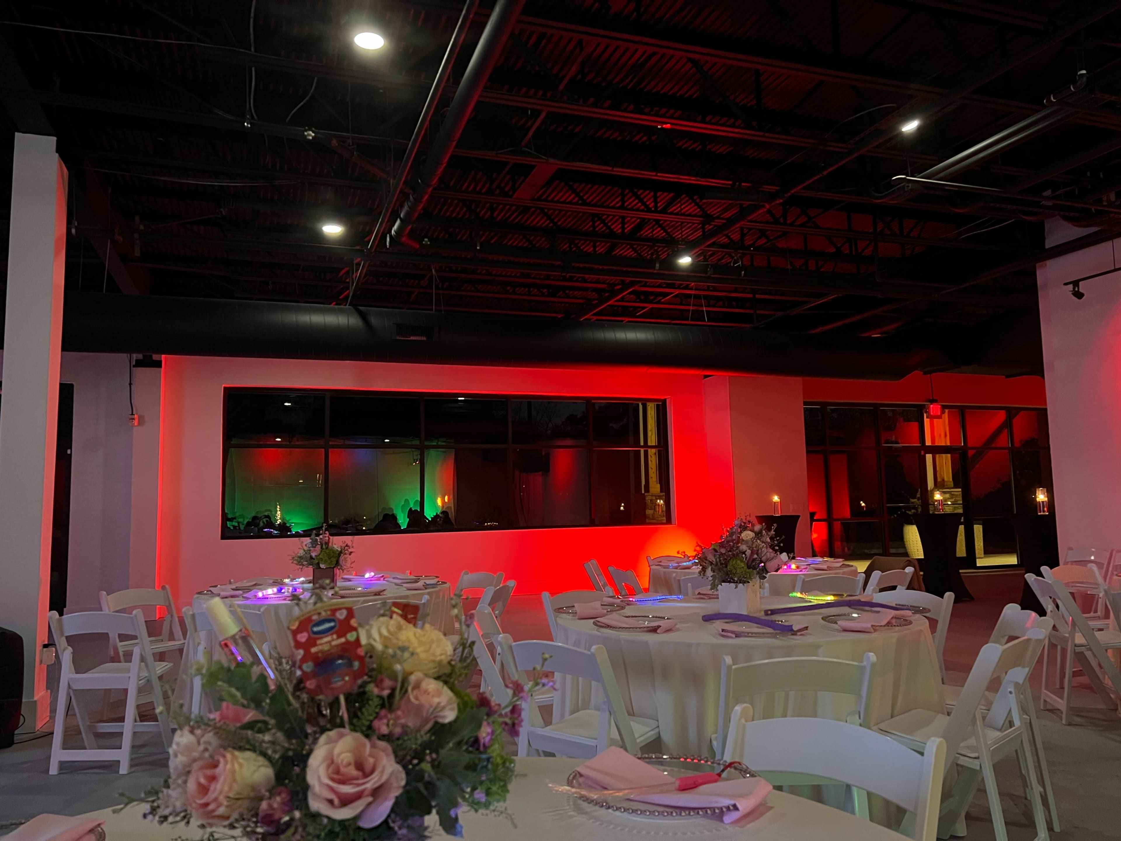 The image shows a dimly lit event space with round tables set for a gathering, colorful lighting on the walls, and large windows revealing guests in the background.