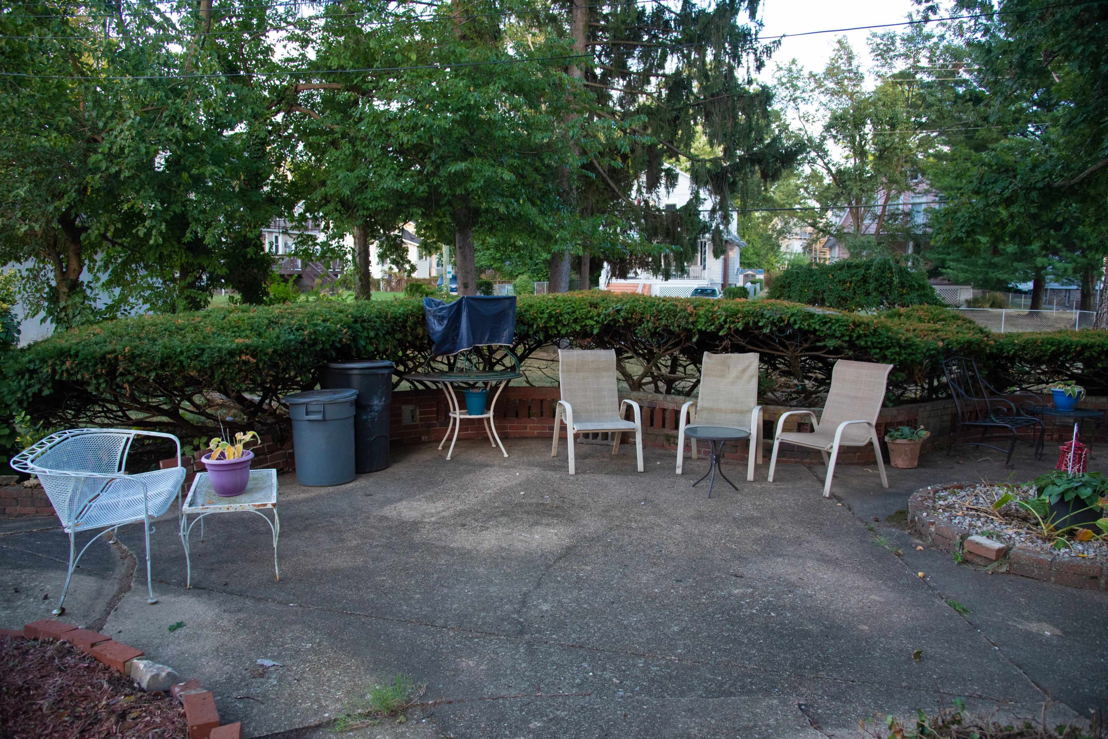 A concrete patio is surrounded by bushes and features several chairs, a trash bin, and potted plants.