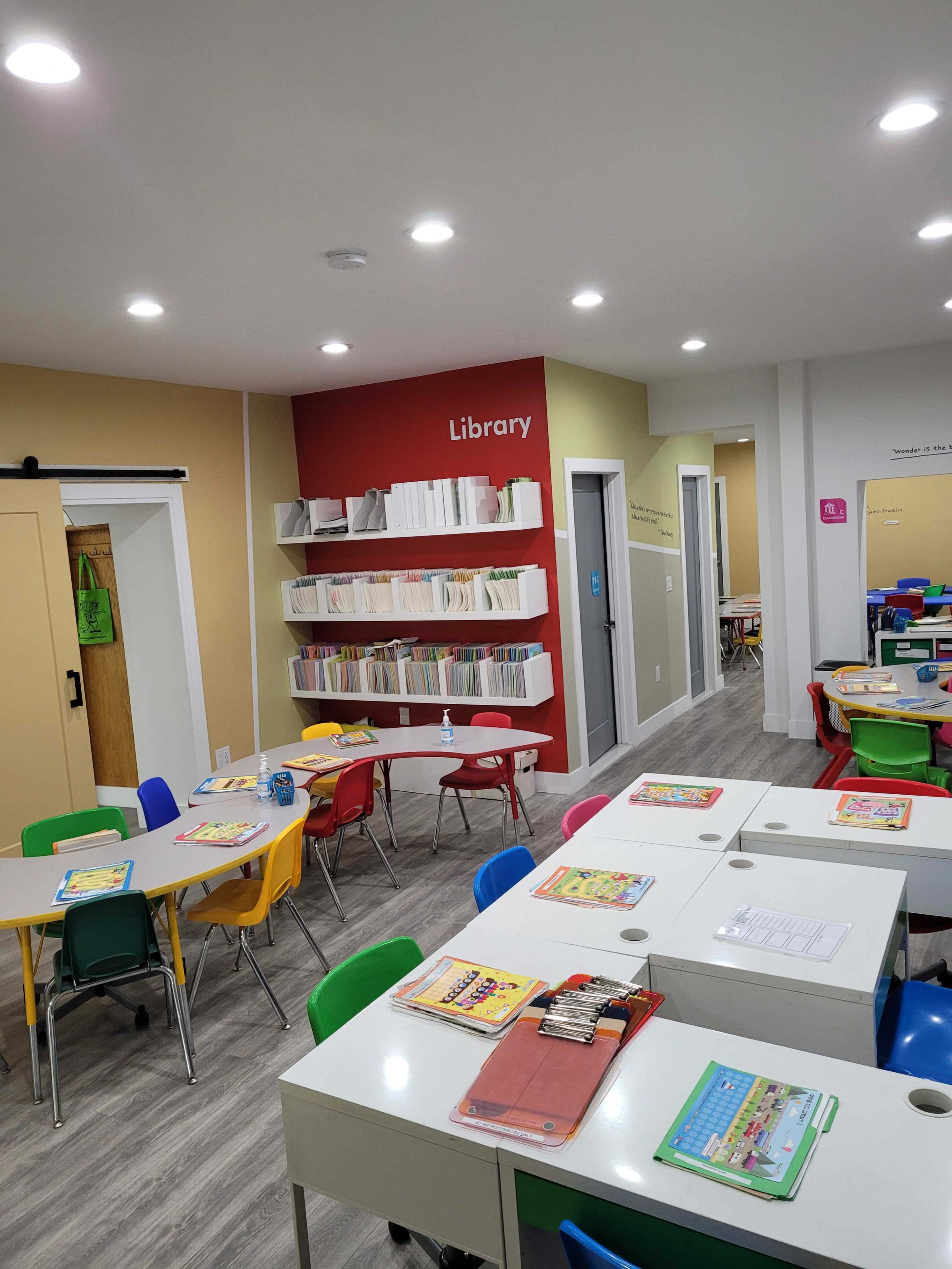 A brightly lit classroom with colorful furniture, including tables and chairs in various colors, and a section labeled "Library" featuring neatly arranged books and materials on shelves.