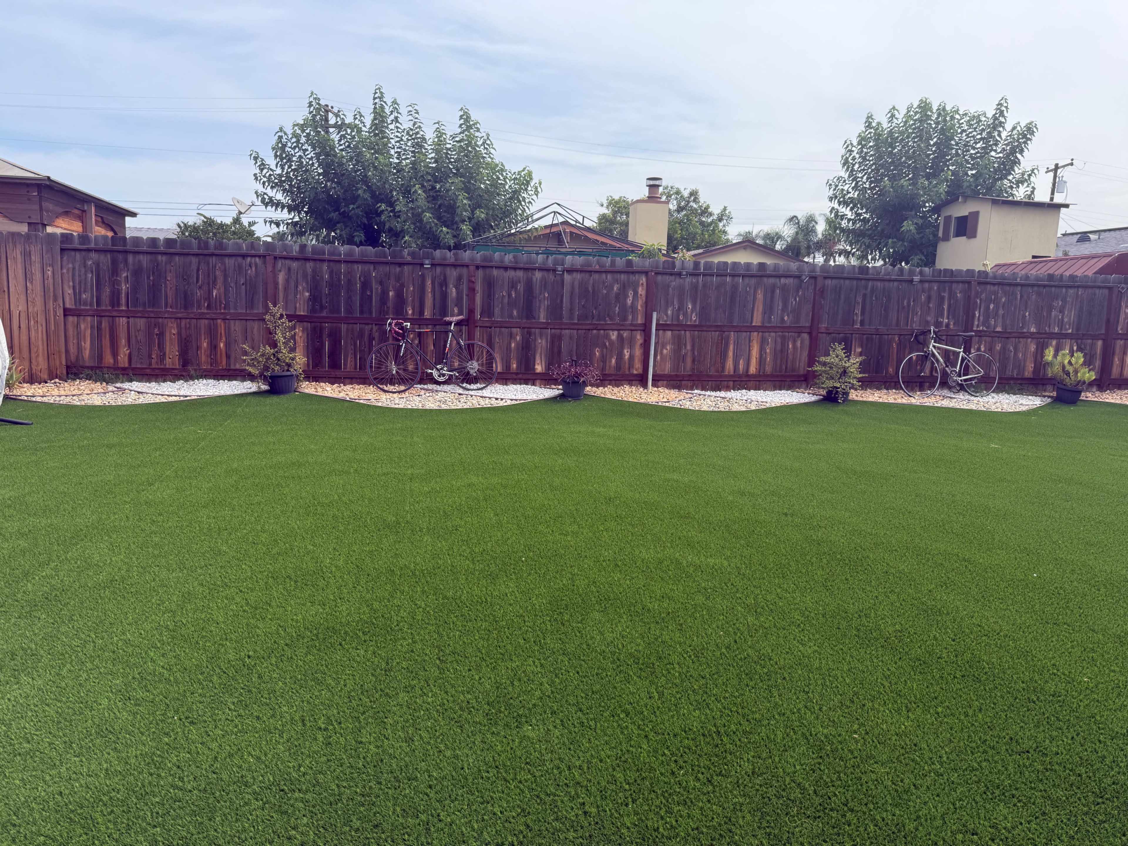 The image shows a green artificial turf yard enclosed by a wooden fence, with two bicycles positioned against the fence and potted plants along the perimeter.