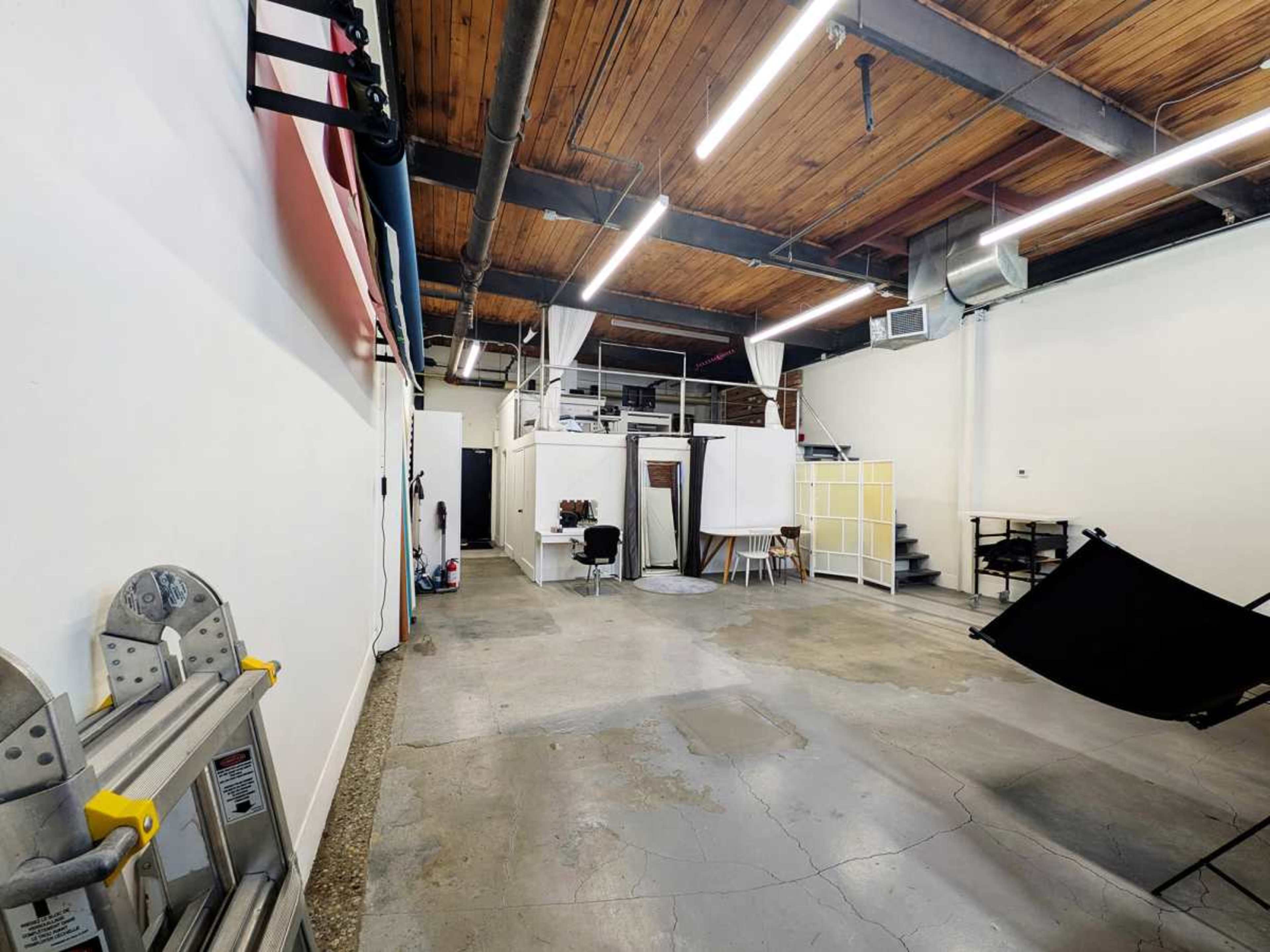 The image shows a spacious, well-lit studio with wooden beams, concrete floors, and various equipment arranged along the walls.