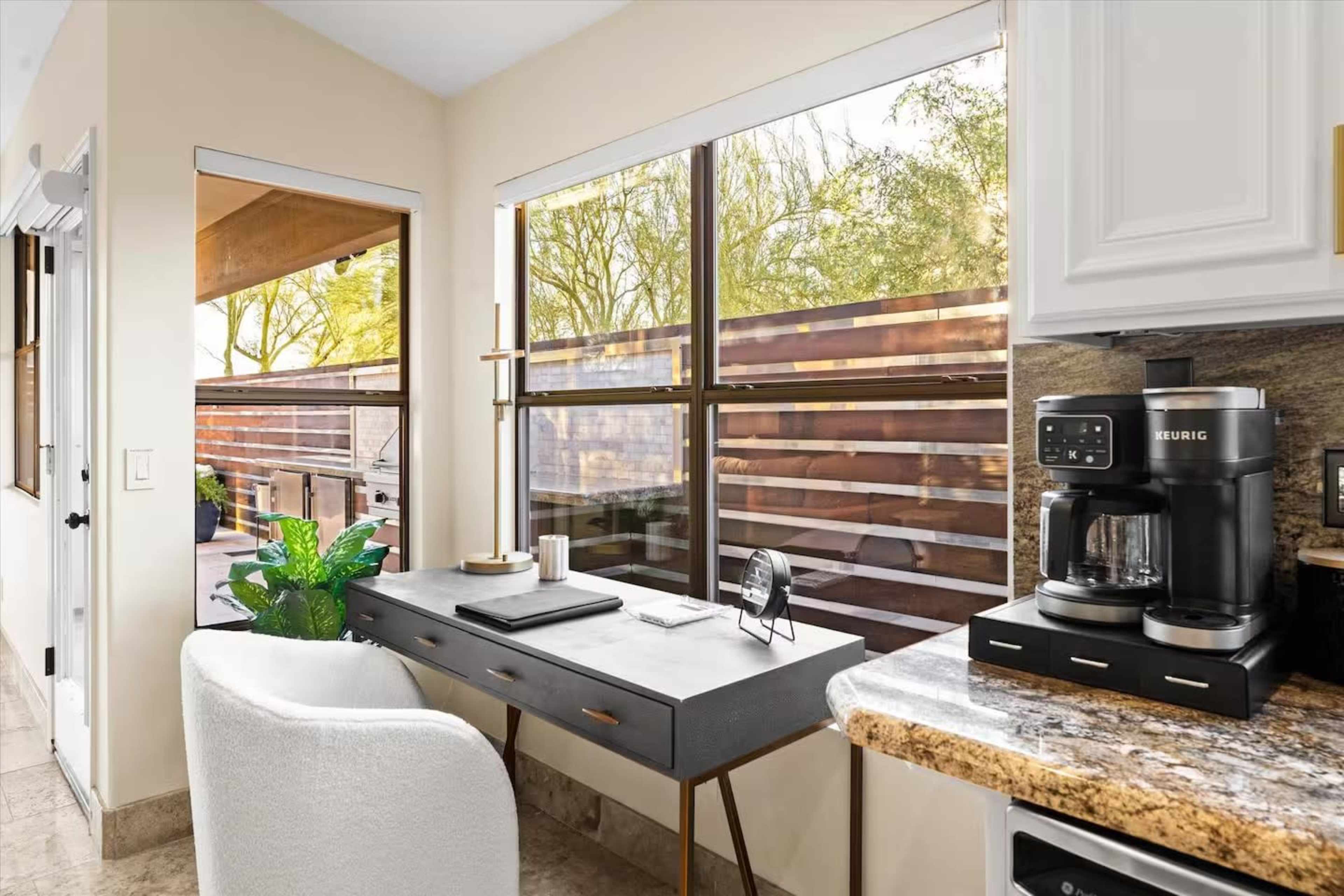 A modern kitchen features a coffee maker, a compact desk with a chair, and large windows overlooking a patio area.