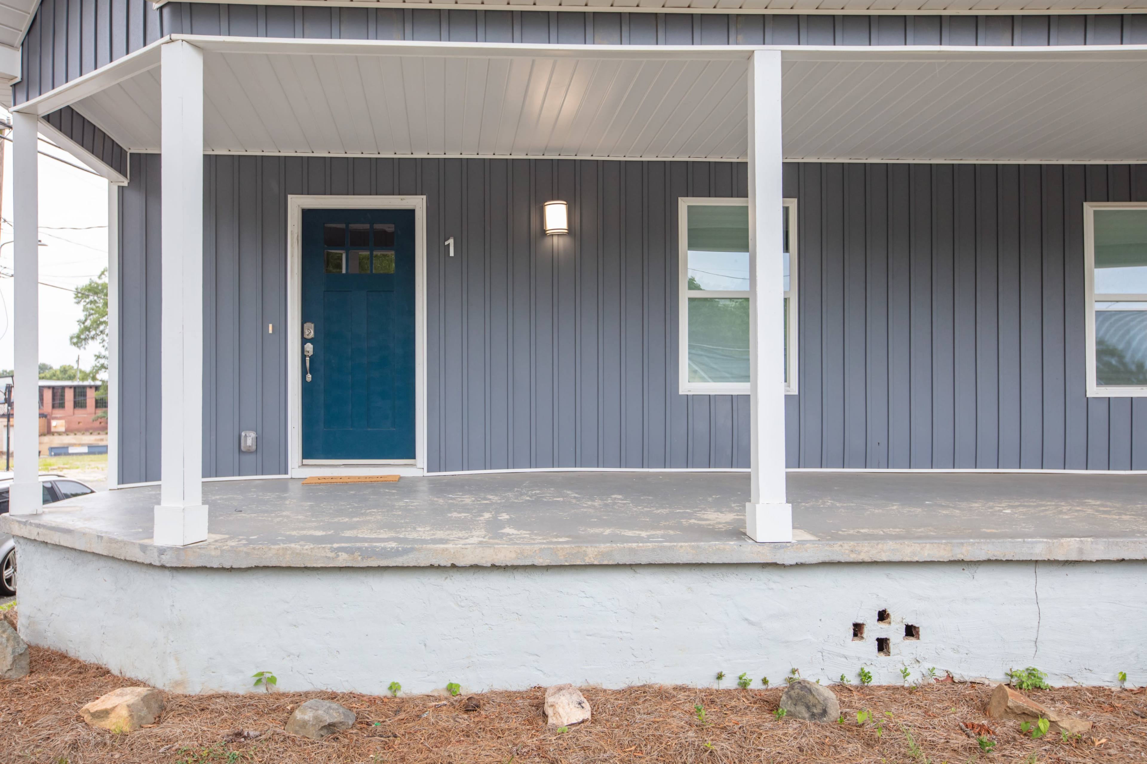 A blue front door on a grey house with a covered porch and a concrete step, surrounded by freshly laid landscaping.