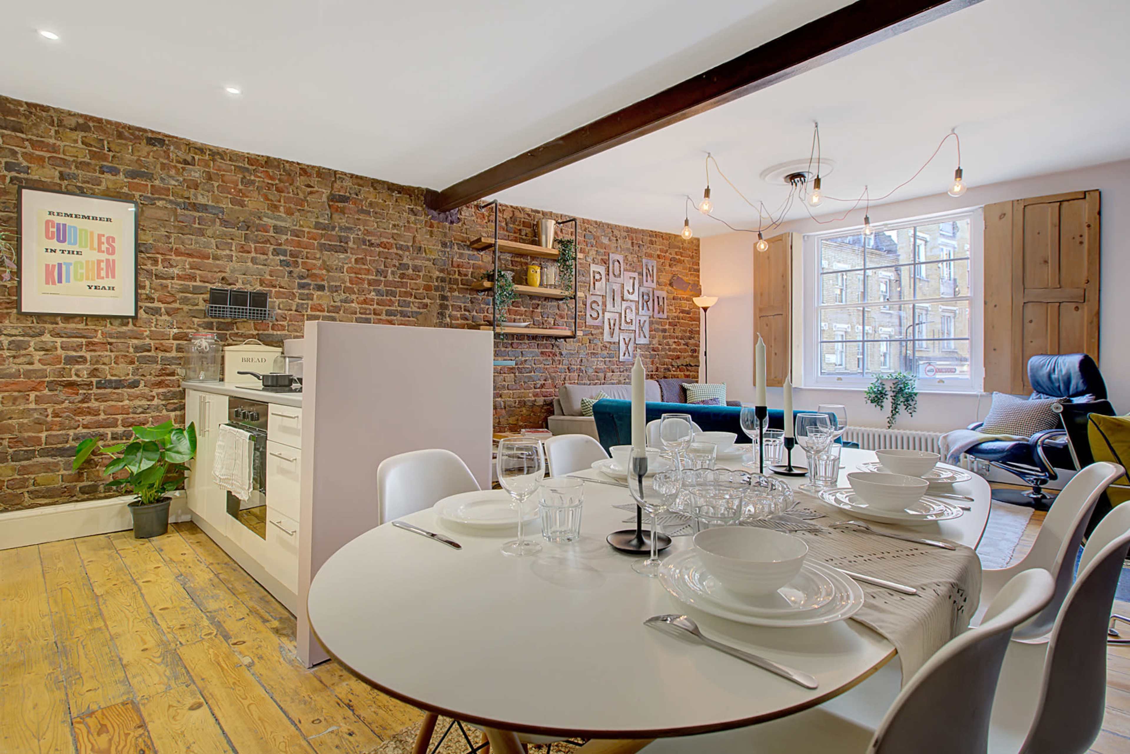 A modern kitchen and dining area features a round table set for a meal, surrounded by white chairs and a backdrop of exposed brick walls.