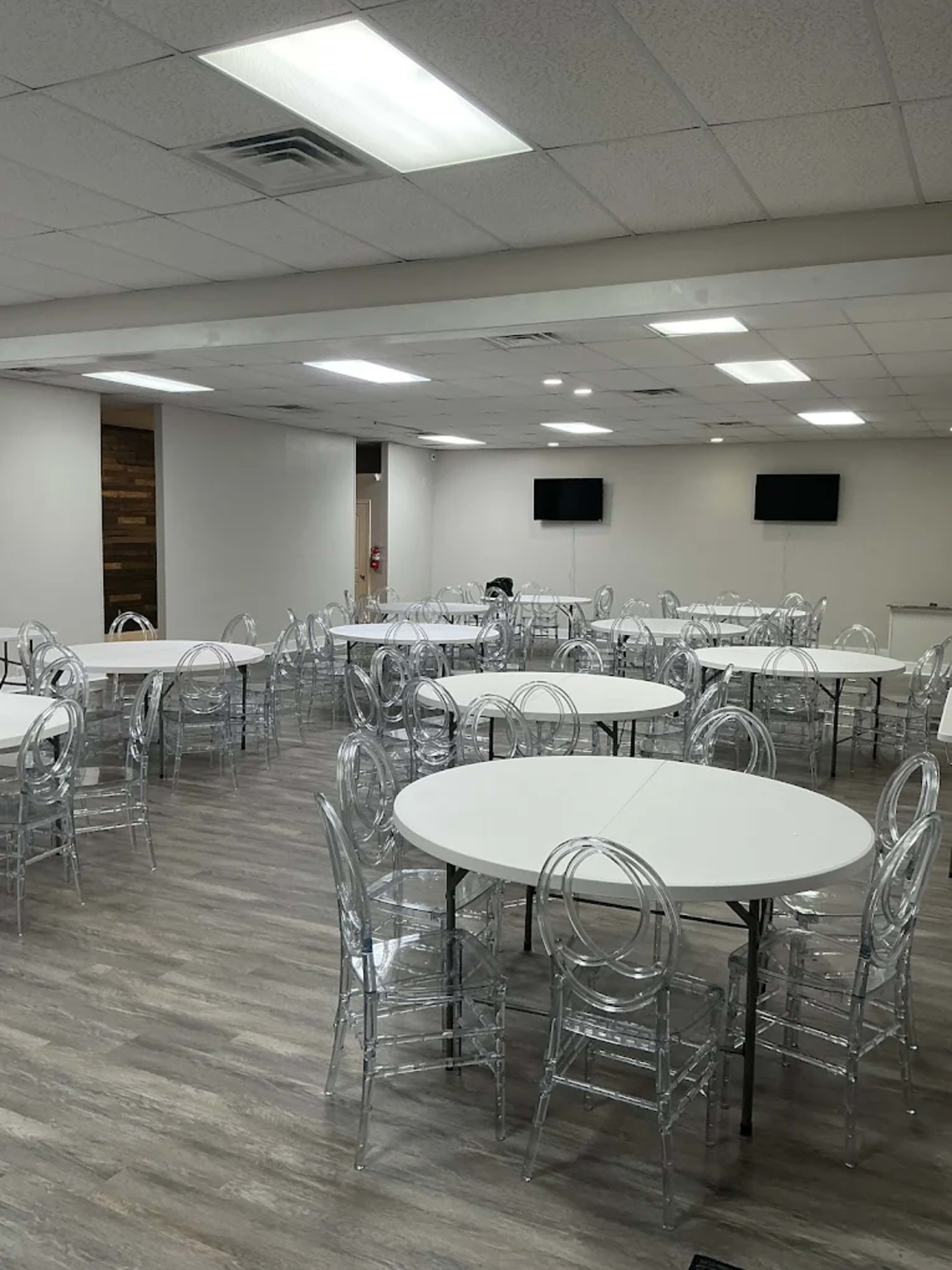 The image shows a spacious event room with multiple round tables surrounded by transparent chairs, all set on a hardwood floor.